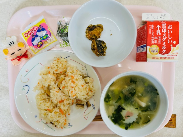 ピンクのトレーに給食が右上から反時計回りで牛乳、さばのごまあげ、ひながし、コック帽をかぶった布製マスコット、ちらしずし、しらたまじると並べられている写真