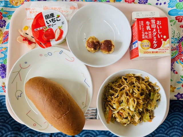 ピンクのトレーに給食が右上から反時計回りで牛乳、あげシューマイ、国産いちごゼリー、コック帽をかぶった布製マスコット、くろパン、ソースやきそばと並べられている写真