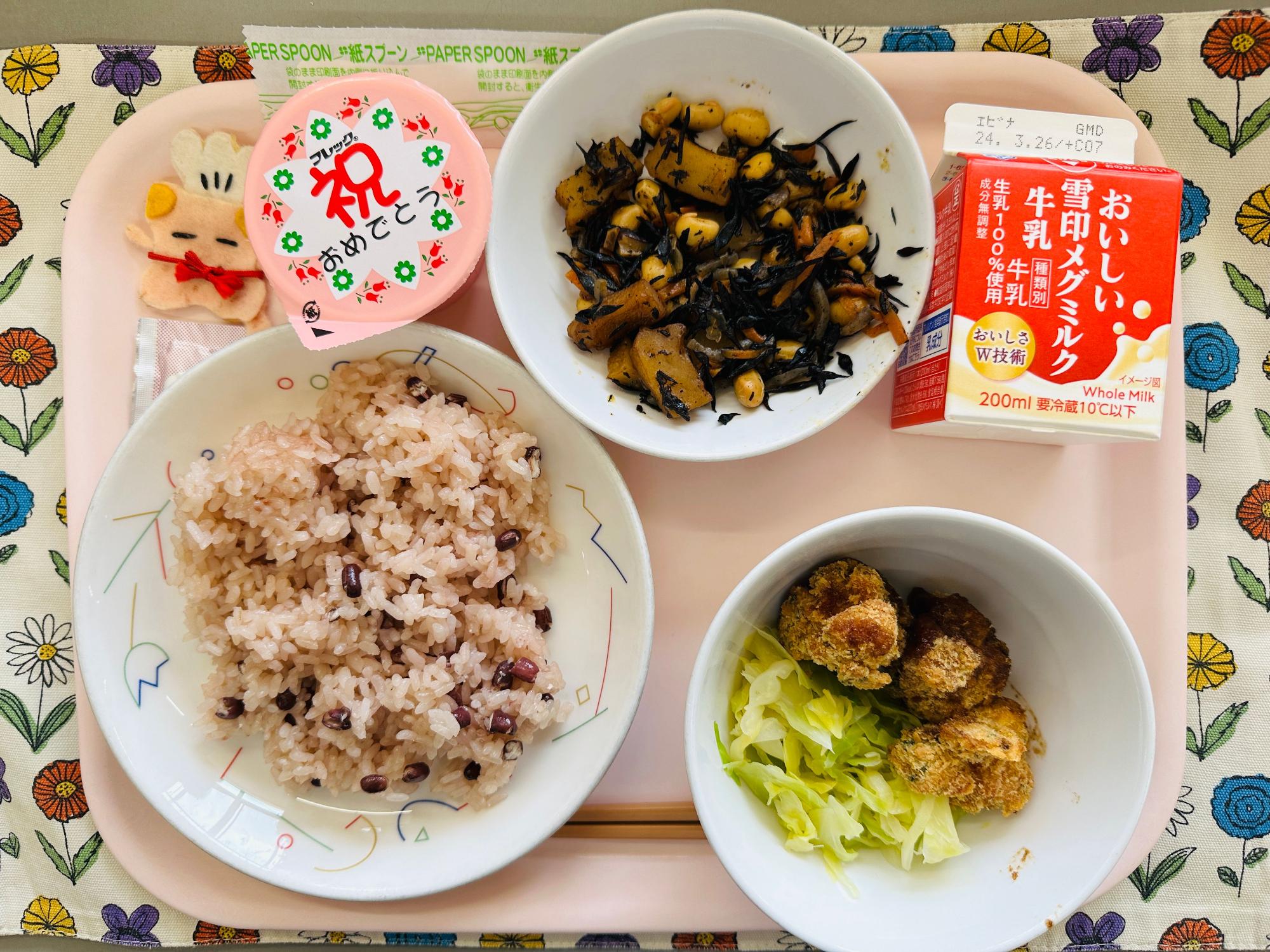 ピンクのトレーに給食が右上から反時計回りで牛乳、ひじきのいりに、デザート、コック帽をかぶった布製マスコット、せきはんとごましお、チキンカツとキャベツソテーと並べられている写真