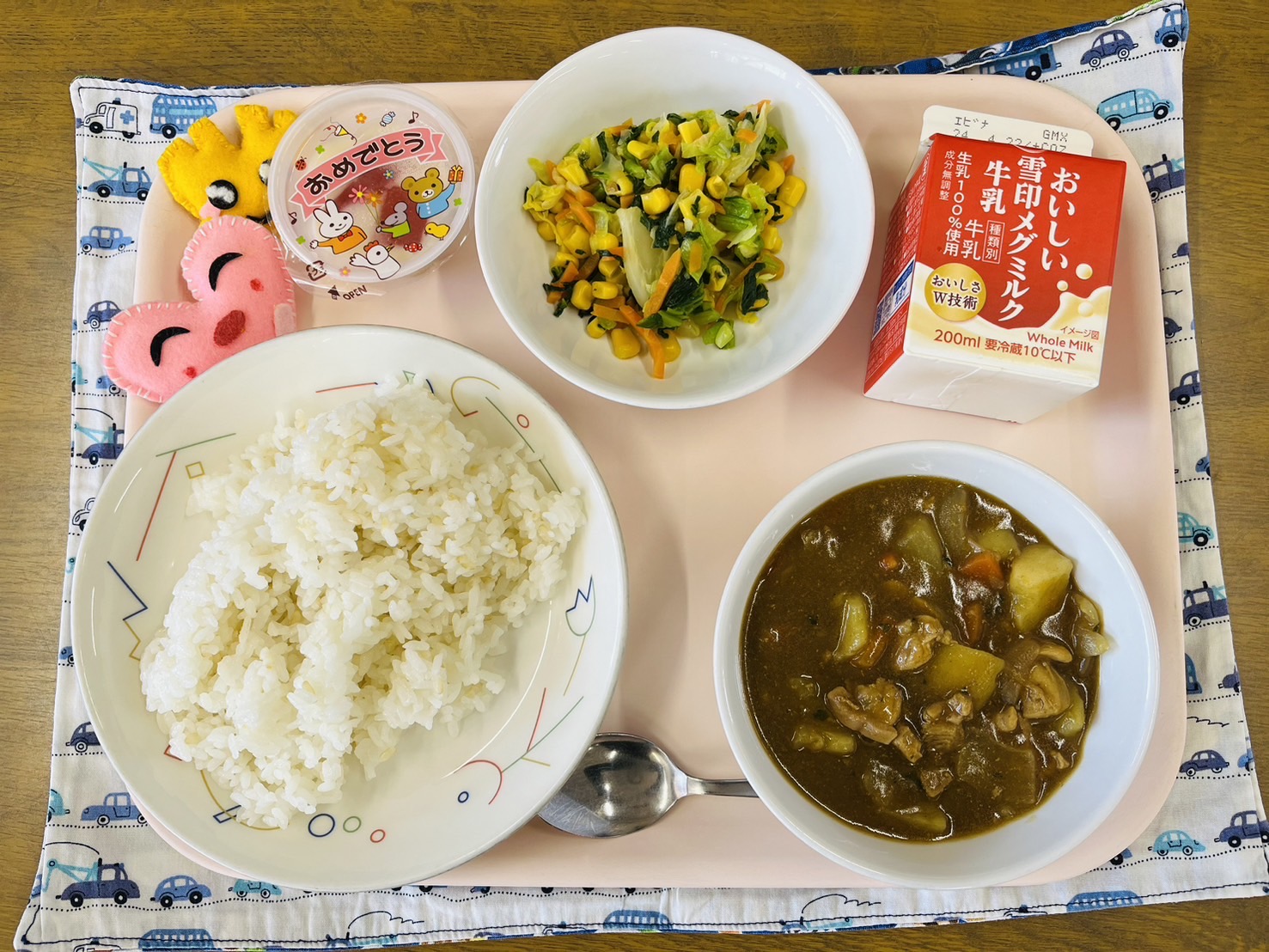 ピンクのトレーに給食が右上から反時計回りで牛乳、やさいソテー、デザート、ハートの形をした黄色とピンク色の布製マスコット、ごはん、チキンカレーと並べられている写真