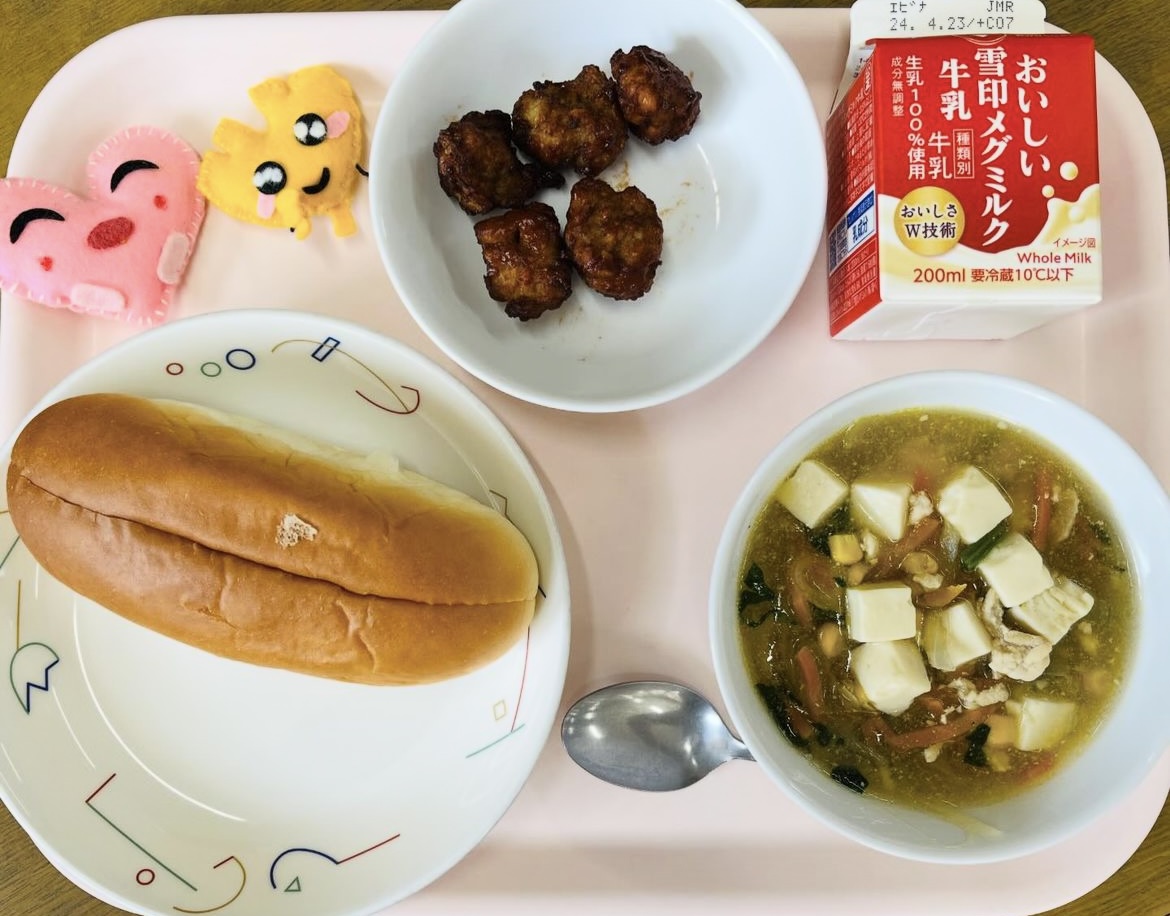 ピンクのトレーに給食が右上から反時計回りで牛乳、ビーンズミートボール、ハートの形をした黄色とピンク色の布製マスコット、あじつけパン、コーン中華スープと並べられている写真