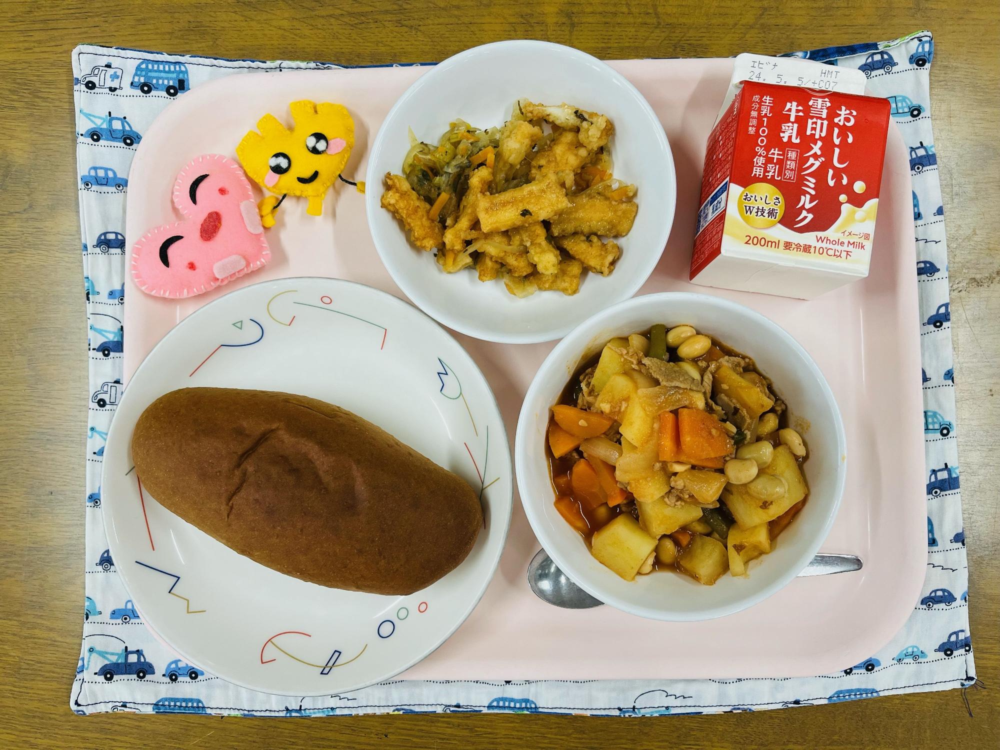 ピンクのトレーに給食が右上から反時計回りで牛乳、いかのマリネ、ハートの形をした黄色とピンク色の布製マスコット、くろパン、ポークビーンズと並べられている写真