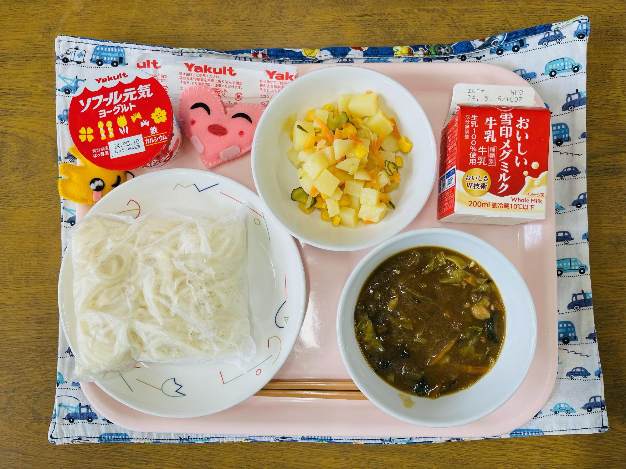 ピンクのトレーに給食が右上から反時計回りで牛乳、ポテトサラダ、デザート、ハートの形をした黄色とピンク色の布製マスコット、ソフトめん、カレーあんかけと並べられている写真