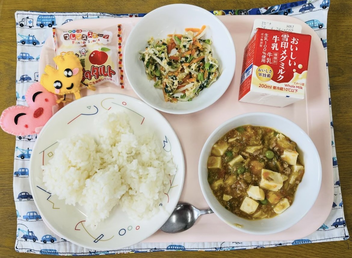 ピンクのトレーに給食が右上から反時計回りで牛乳、鶏肉と野菜のナムル、デザート、ハートの形をした黄色とピンク色の布製マスコット、ごはん、マーボー豆腐と並べられている写真