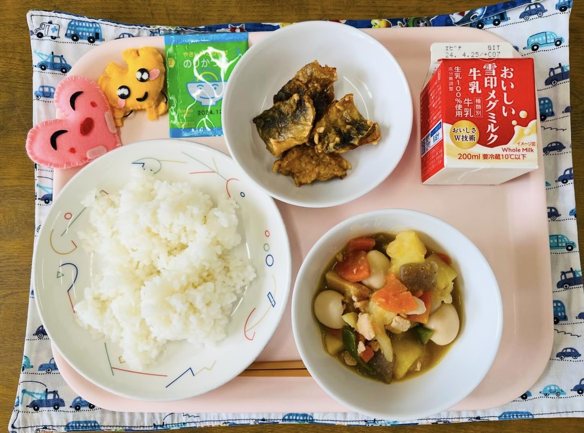 ピンクのトレーに給食が右上から反時計回りで牛乳、あじのからあげ、ふりかけ、ハートの形をした黄色とピンク色の布製マスコット、ごはん、春野菜のうま煮と並べられている写真