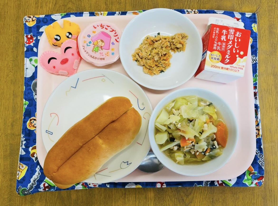 ピンクのトレーに給食が右上から反時計回りで牛乳、ツナサンドの具、デザート、ハートの形をした黄色とピンク色の布製マスコット、サンドパン、野菜のスープ煮と並べられている写真