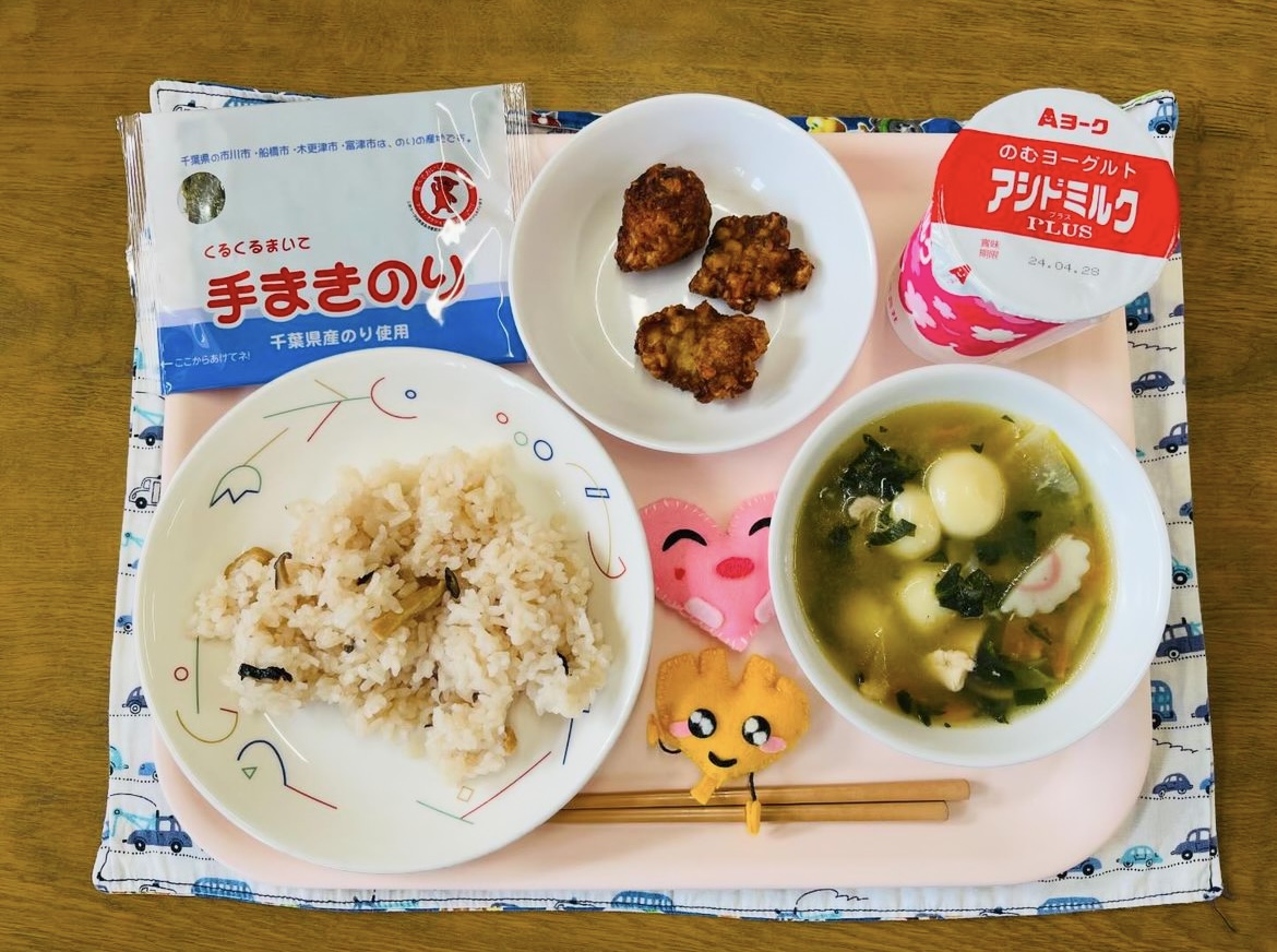 ピンクのトレーに給食が右上から反時計回りで発酵乳、豚肉のふうみあげ、てまきのり、ハートの形をした黄色とピンク色の布製マスコット、さくらずし、しらたまじると並べられている写真