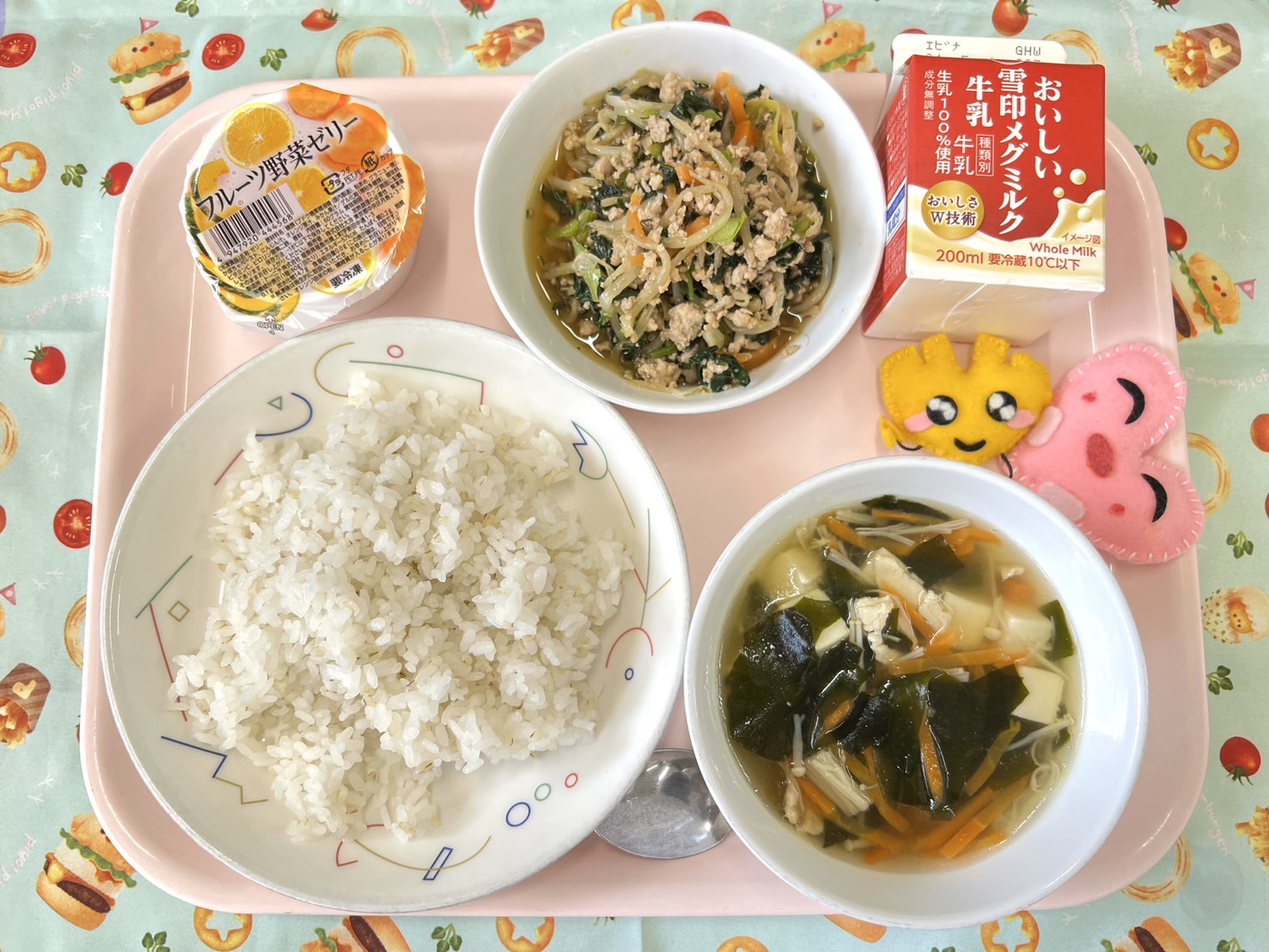 ピンクのトレーに給食が右上から反時計回りで牛乳、ビビンバ、デザート、むぎいりごはん、わかめスープ、ハートの形をした黄色とピンク色の布製マスコットと並べられている写真