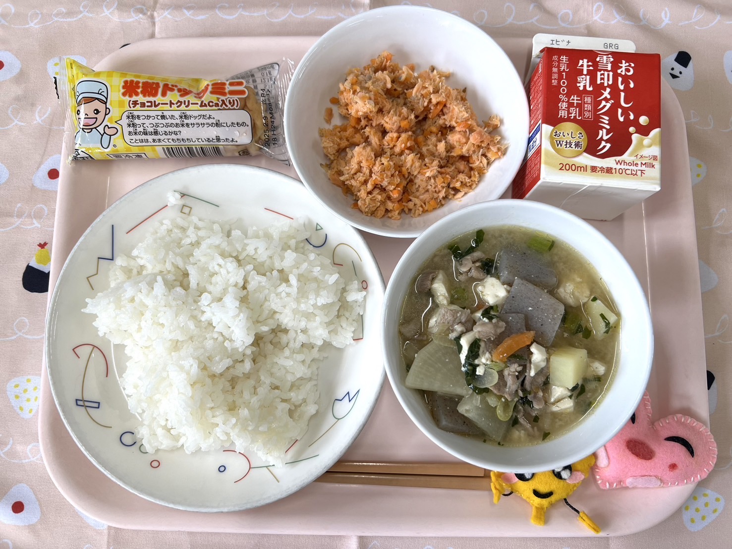 ピンクのトレーに給食が右上から反時計回りで牛乳、さけそぼろ、デザート、ごはん、豚汁、ハートの形をした黄色とピンク色の布製マスコットと並べられている写真