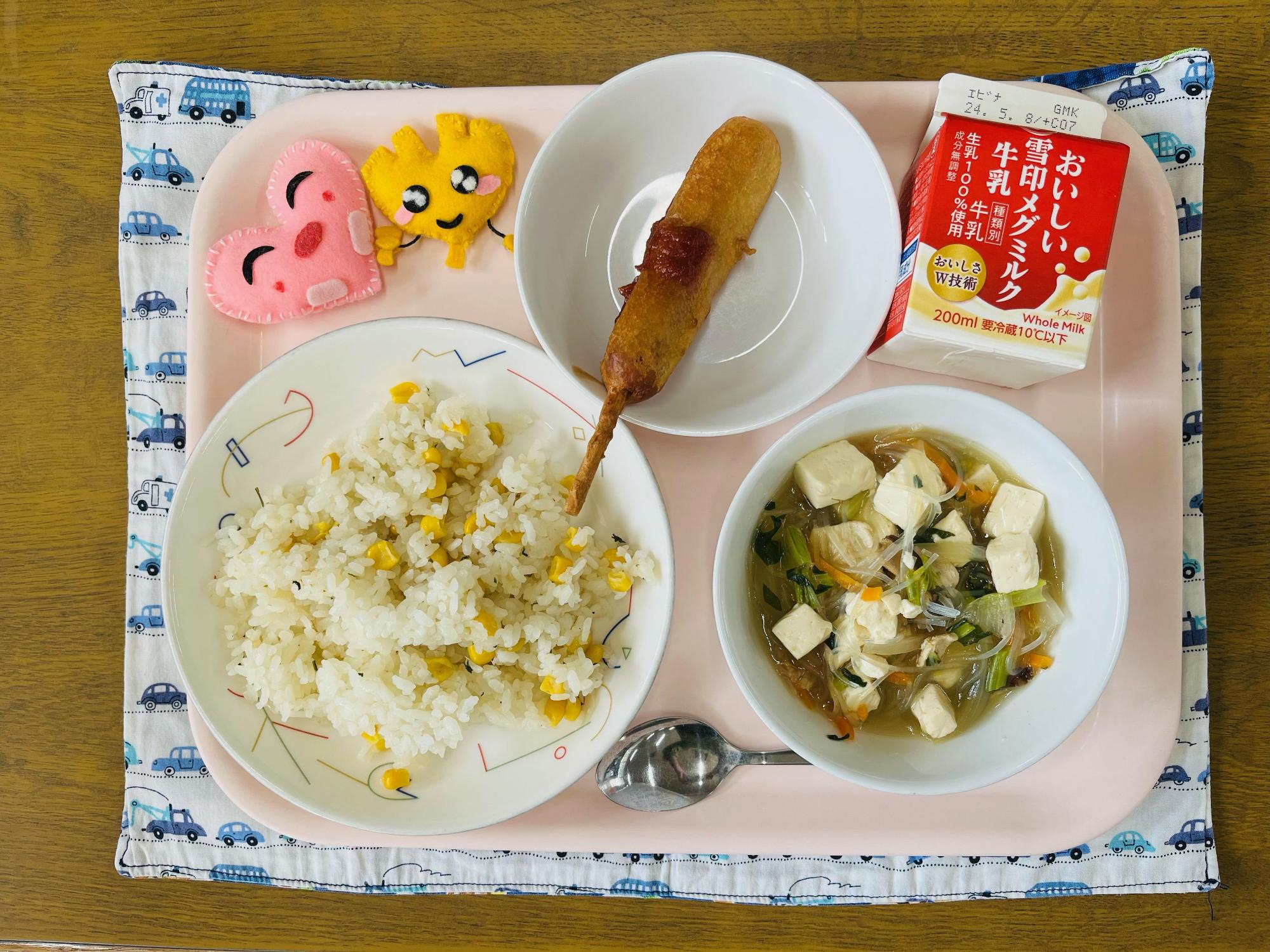 ピンクのトレーに給食が右上から反時計回りで牛乳、アメリカンドッグ、ハートの形をした黄色とピンク色の布製マスコット、コーンピラフ、はるさめスープと並べられている写真