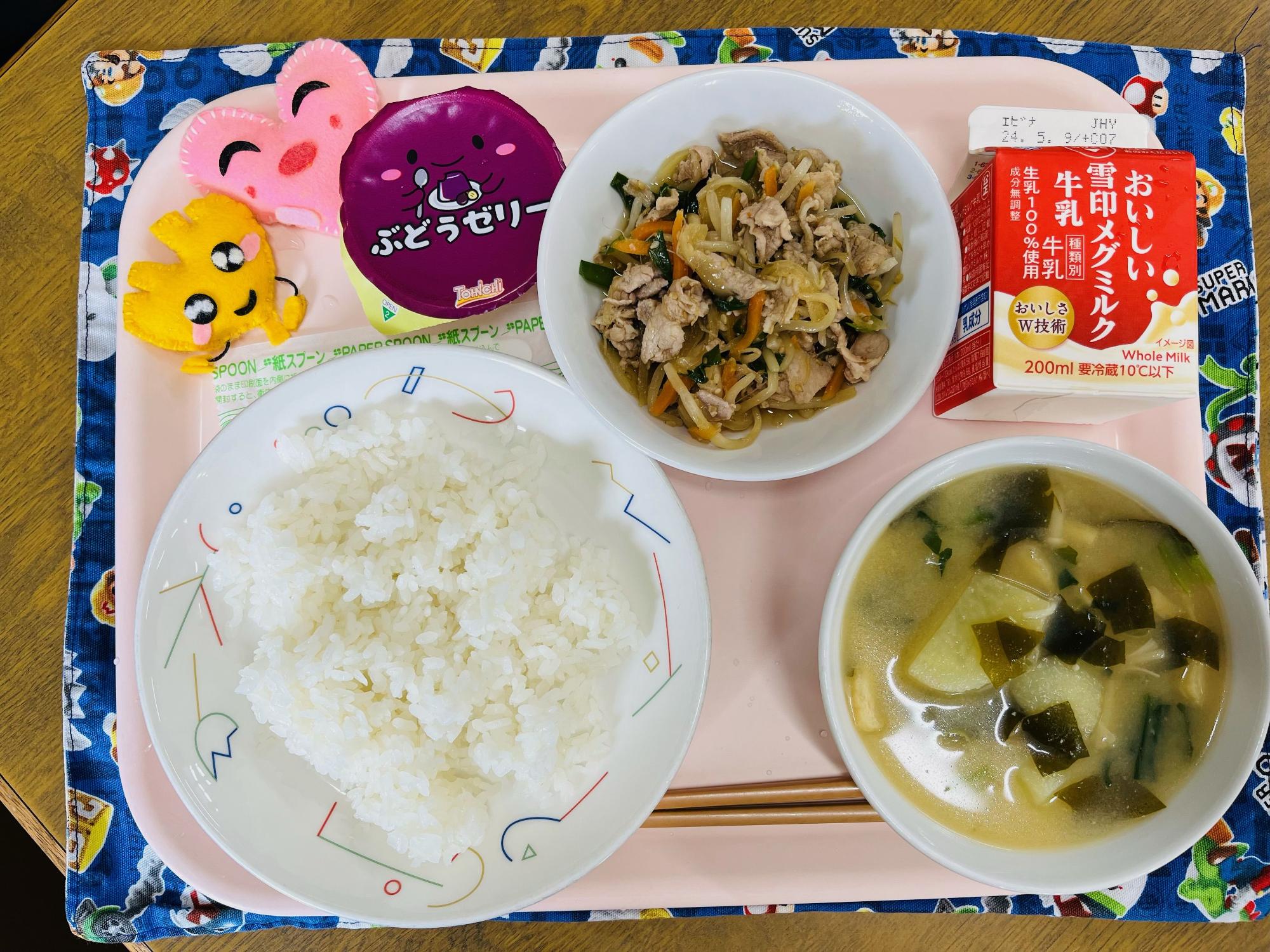 ピンクのトレーに給食が右上から反時計回りで牛乳、野菜入りやきにく、デザート、ハートの形をした黄色とピンク色の布製マスコット、ごはん、みそしると並べられている写真