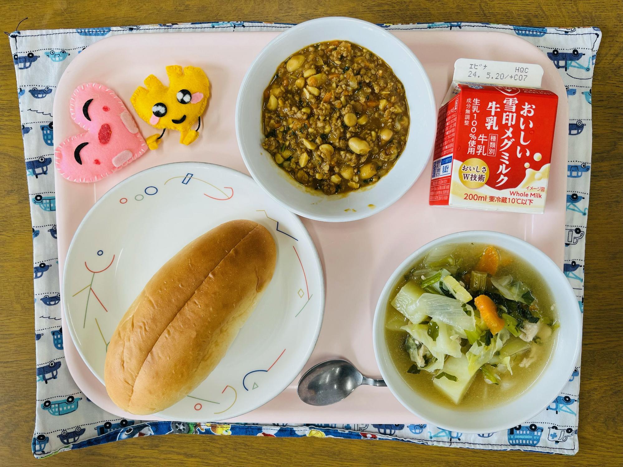 ピンクのトレーに給食が右上から反時計回りで牛乳、ドライカレー、ハートの形をした黄色とピンク色の布製マスコット、味付けパン、野菜のスープ煮と並べられている写真