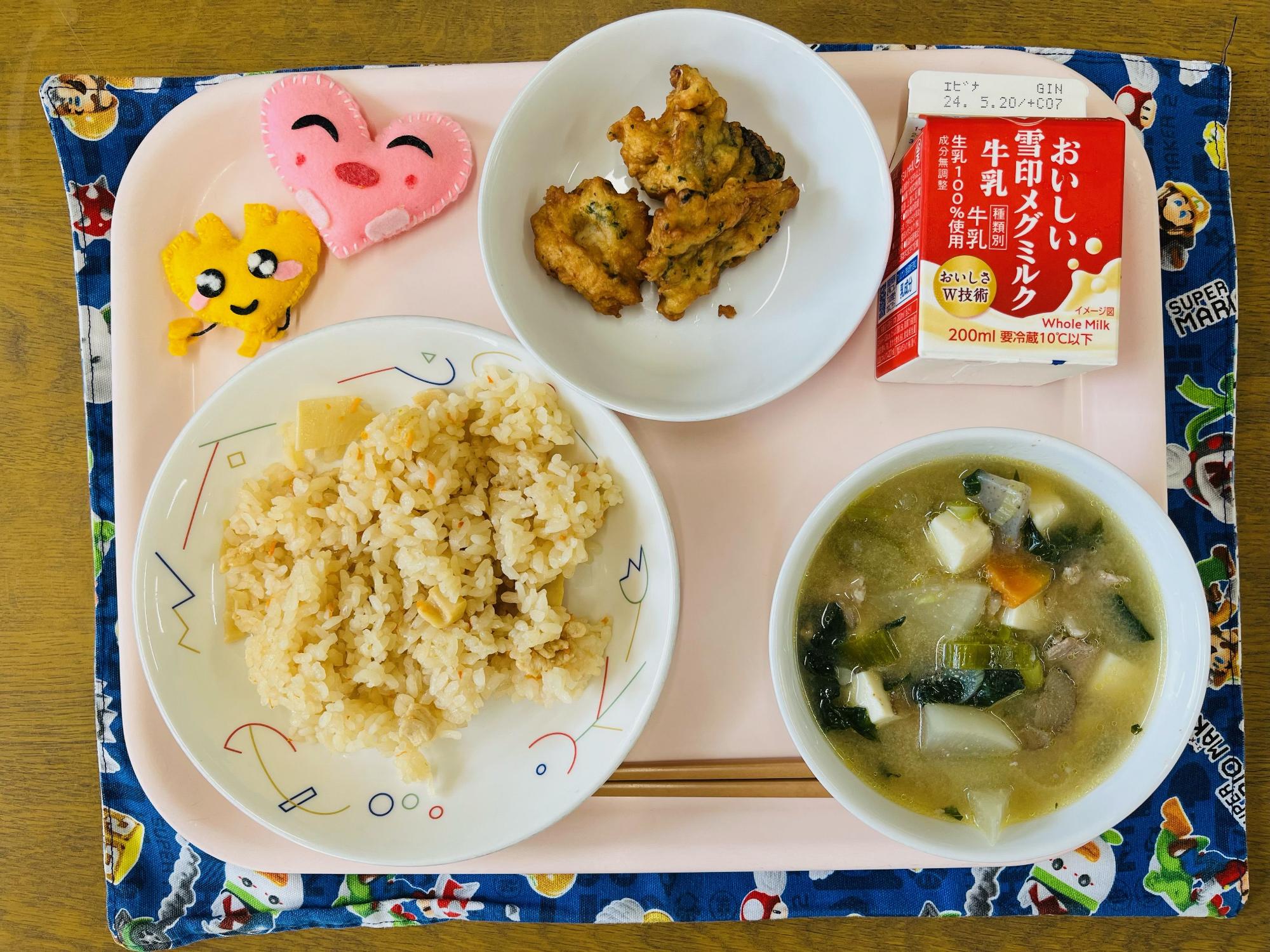 ピンクのトレーに給食が右上から反時計回りで牛乳、あじのふわっと揚げ、ハートの形をした黄色とピンク色の布製マスコット、たけのこごはん、あっさり豚汁と並べられている写真