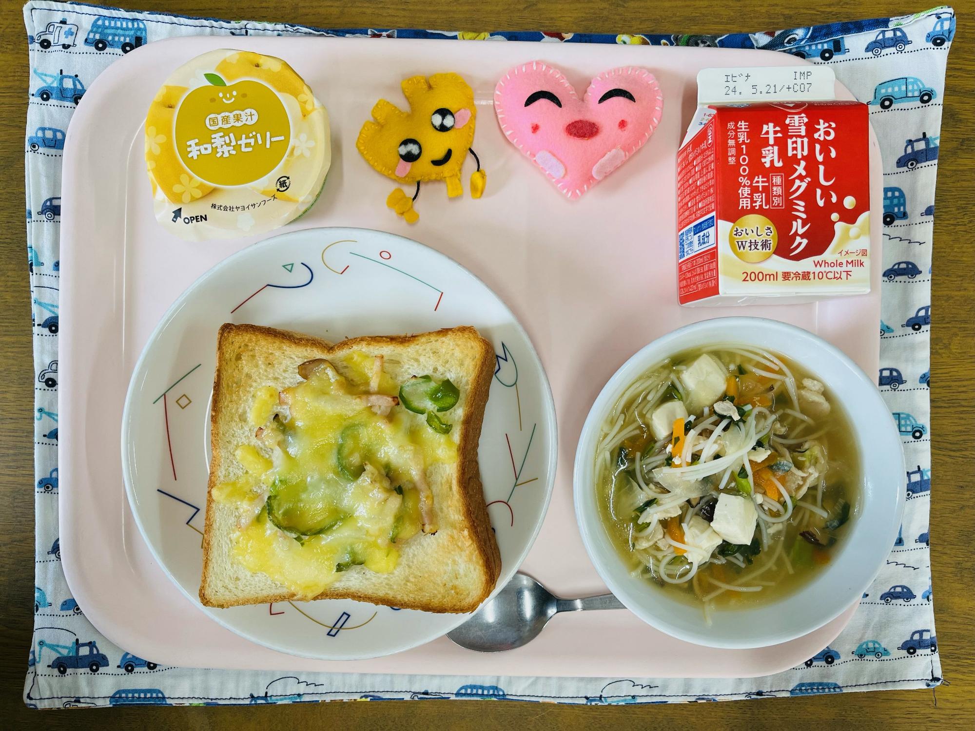 ピンクのトレーに給食が右上から反時計回りで牛乳、ハートの形をした黄色とピンク色の布製マスコット、デザート、ピザトースト、ビーフンスープと並べられている写真