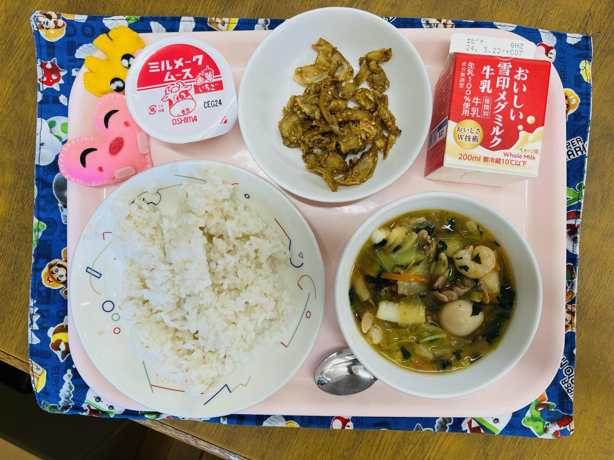 ピンクのトレーに給食が右上から反時計回りで牛乳、ごぼうの甘辛だれからめ、デザート、ハートの形をした黄色とピンク色の布製マスコット、むぎいりご飯、八宝菜と並べられている写真