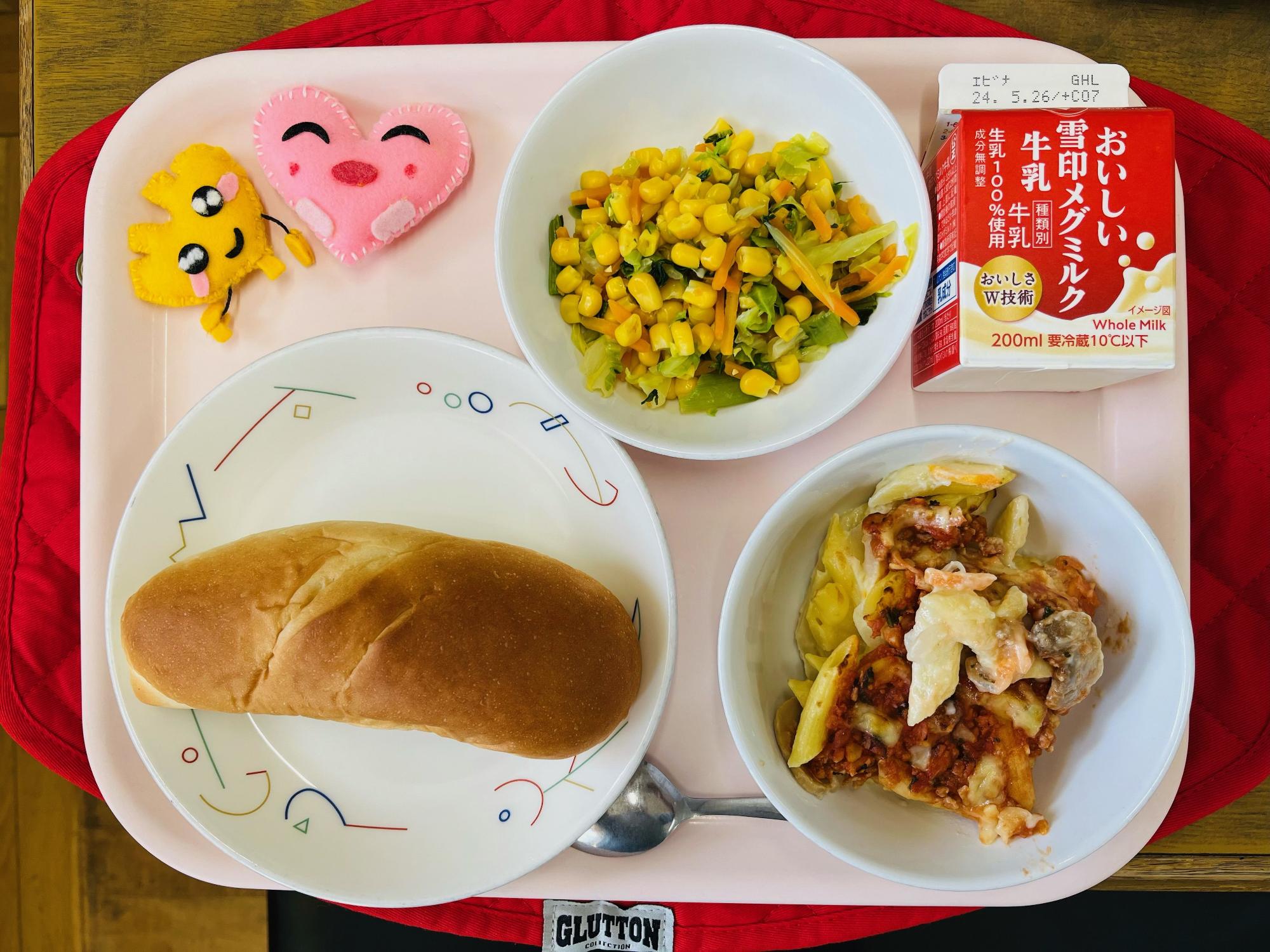 ピンクのトレーに給食が右上から反時計回りで牛乳、コーンソテー、ハートの形をした黄色とピンク色の布製マスコット、ミルクパン、ソイミートグラタンと並べられている写真