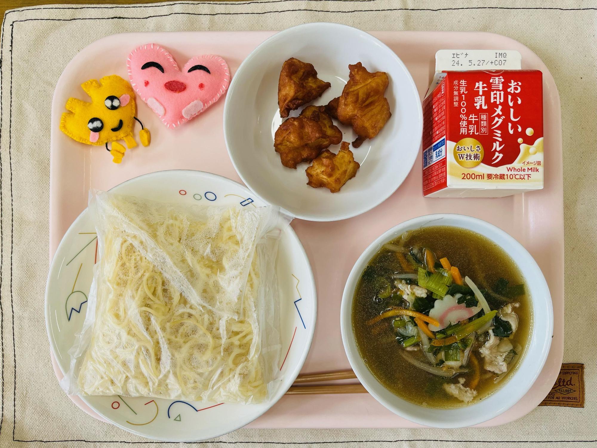 ピンクのトレーに給食が右上から反時計回りで牛乳、じゃがじゃが、ハートの形をした黄色とピンク色の布製マスコット、麺、しょうゆラーメンのスープと並べられている写真
