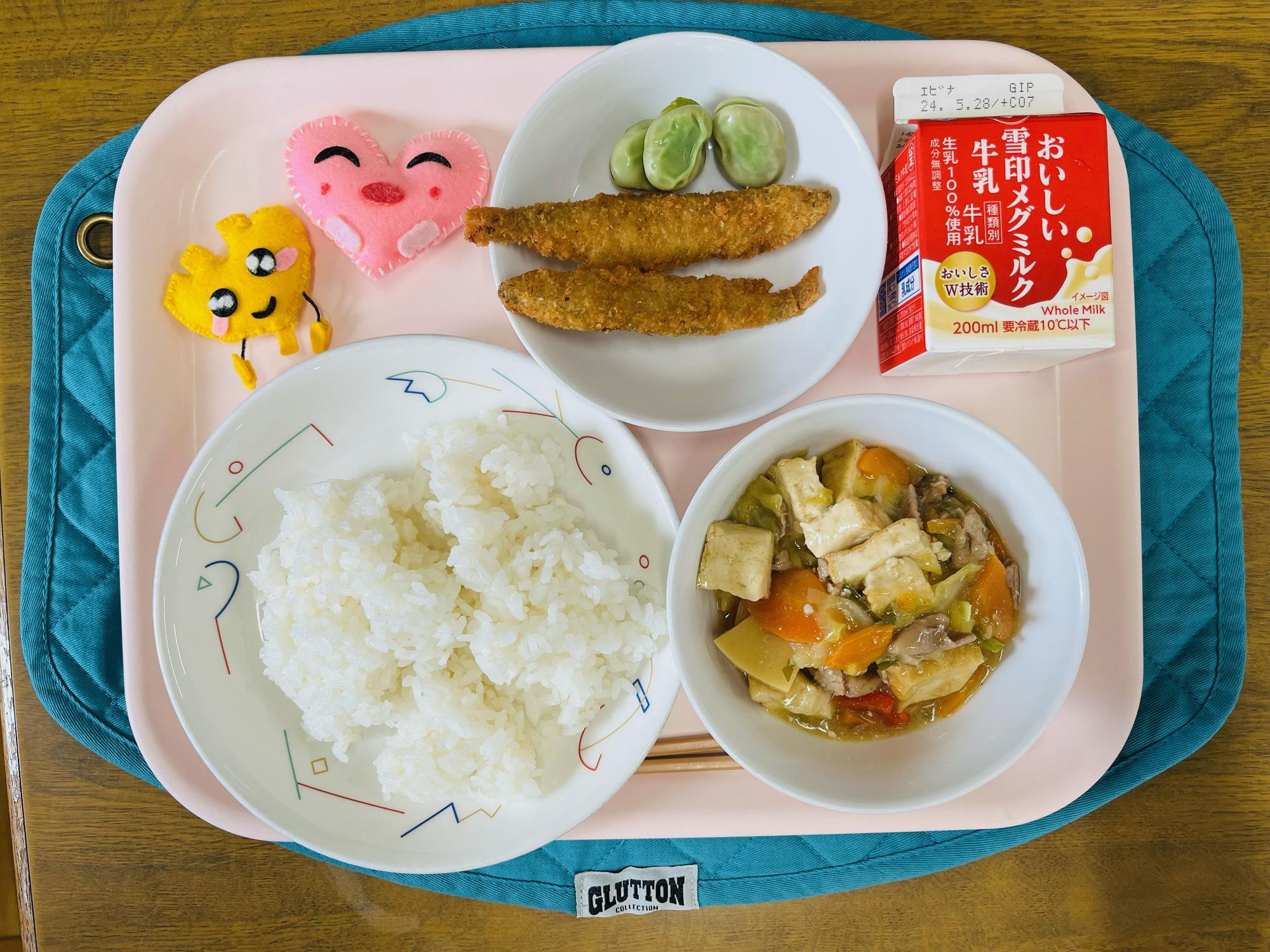 ピンクのトレーに給食が右上から反時計回りで牛乳、そらまめとししゃもフライ、ハートの形をした黄色とピンク色の布製マスコット、ごはん、ジャージャン豆腐と並べられている写真