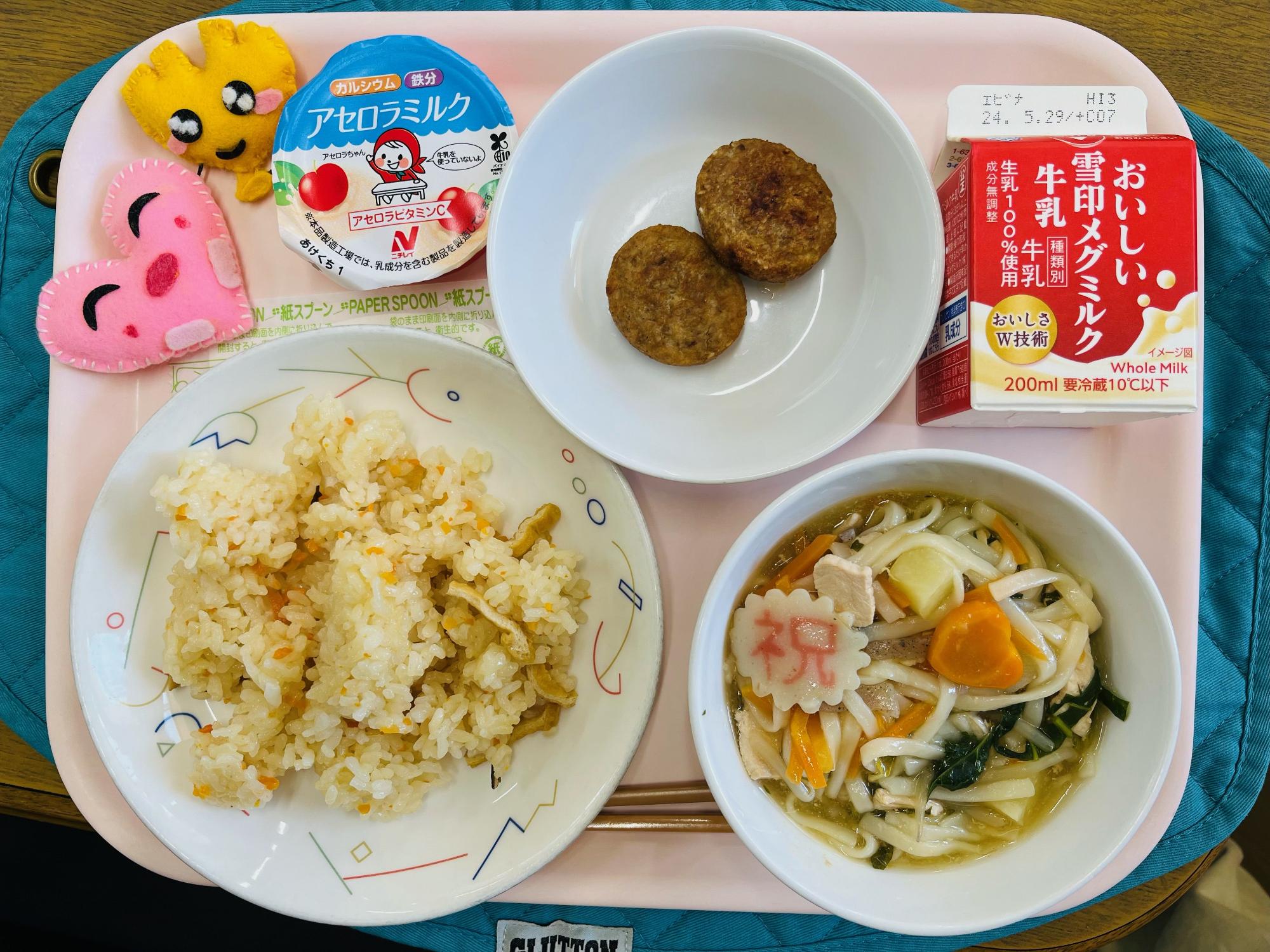 ピンクのトレーに給食が右上から反時計回りで牛乳、白ごまつくね、デザート、ハートの形をした黄色とピンク色の布製マスコット、五目ずし、あんかけ汁と並べられている写真