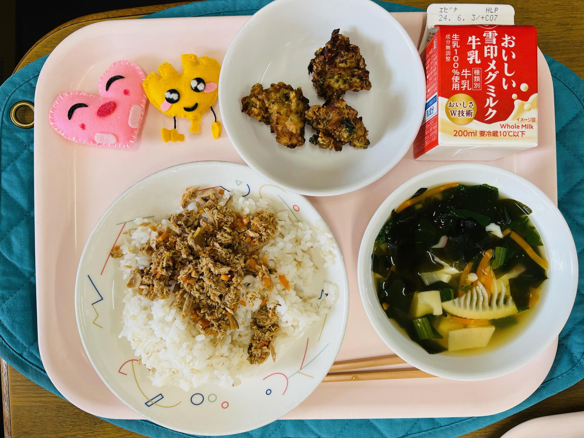 ピンクのトレーに給食が右上から反時計回りで牛乳、とりにくのころも揚げ、ハートの形をした黄色とピンク色の布製マスコット、かつおそぼろご飯、わかたけ汁と並べられている写真