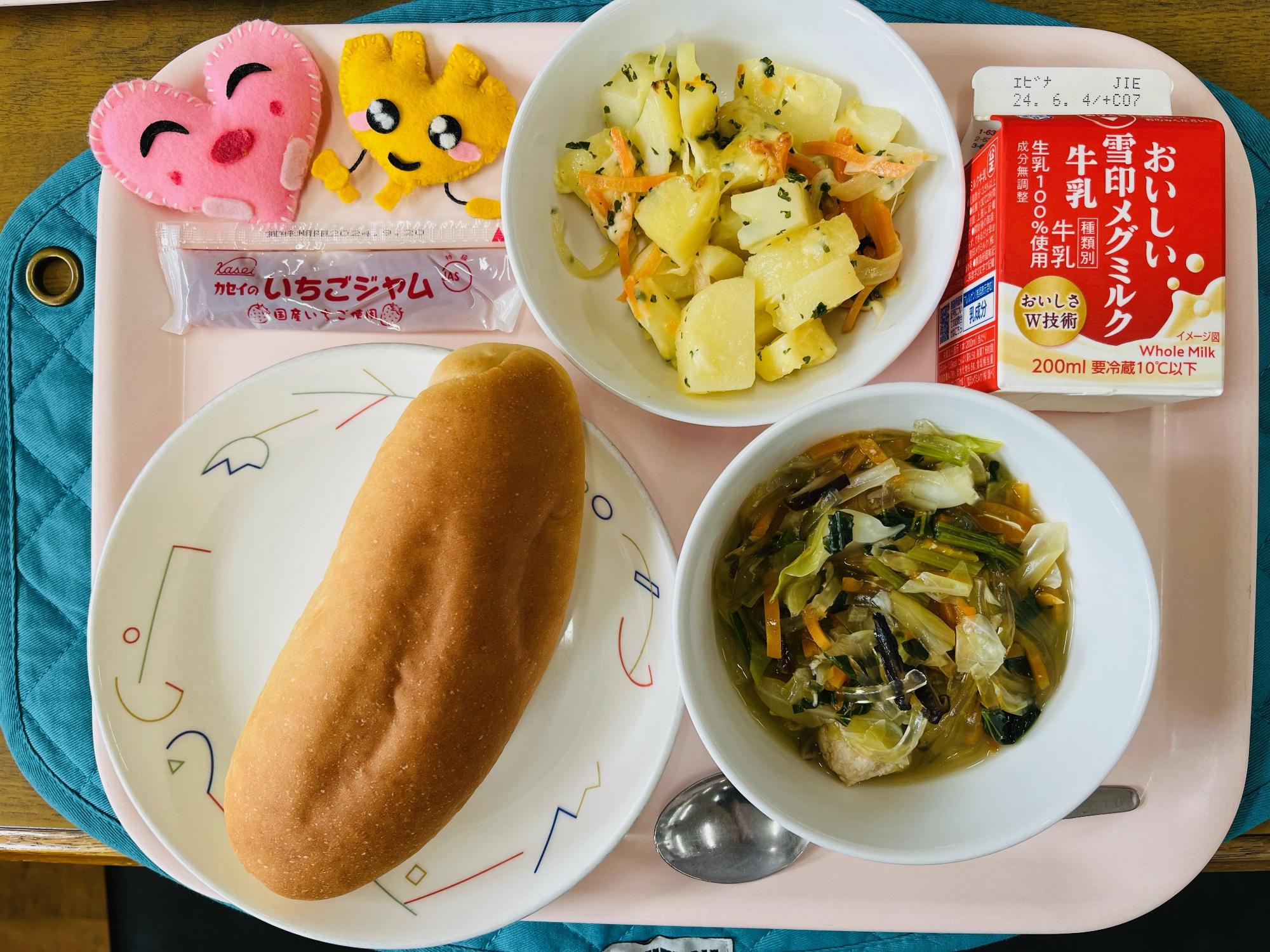 ピンクのトレーに給食が右上から反時計回りで牛乳、じゃがいものチーズ焼き、ジャム、ハートの形をした黄色とピンク色の布製マスコット、コッペパン、とりだんごとキャベツのスープと並べられている写真