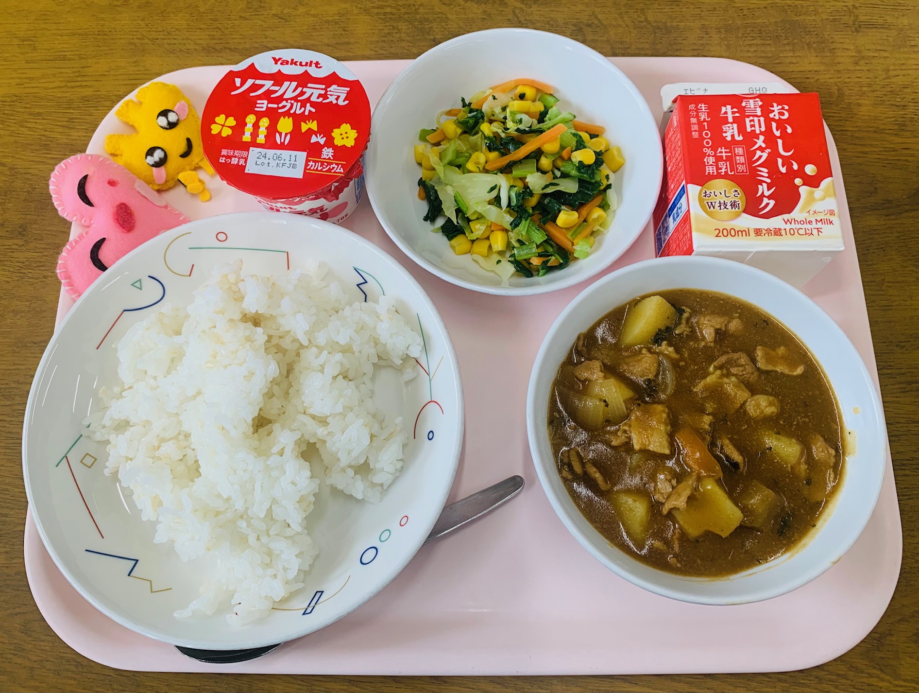 ピンクのトレーに給食が右上から反時計回りで牛乳、野菜ソテー、デザート、ハートの形をした黄色とピンク色の布製マスコット、ごはん、カレーと並べられている写真