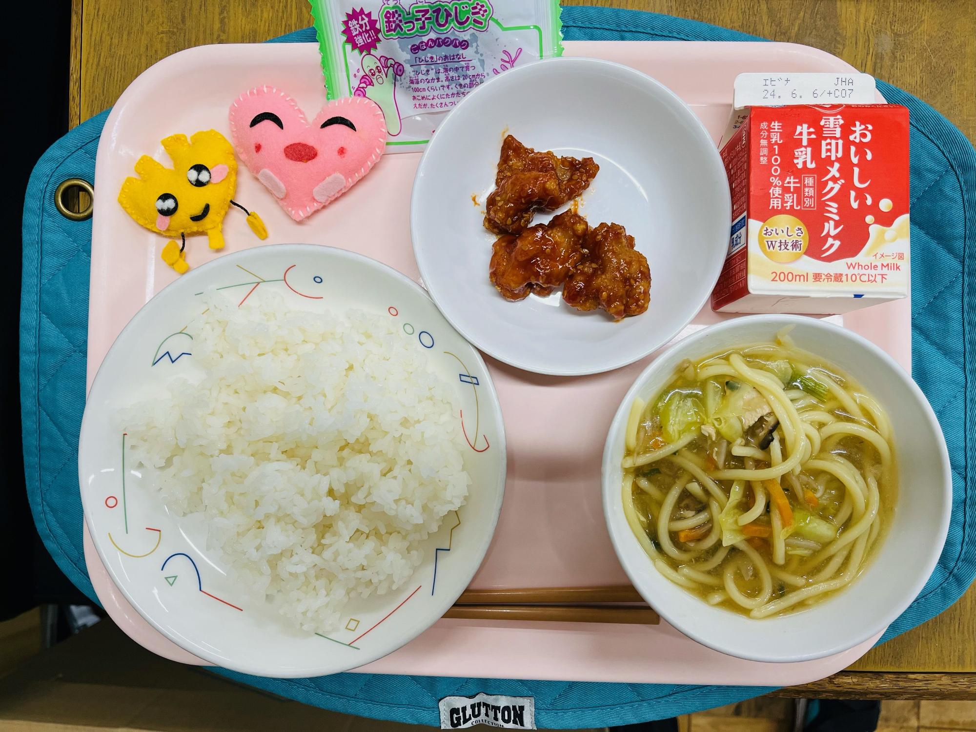 ピンクのトレーに給食が右上から反時計回りで牛乳、ヤンニョムチキン、佃煮、ハートの形をした黄色とピンク色の布製マスコット、ごはん、ちゃんぽんスープと並べられている写真