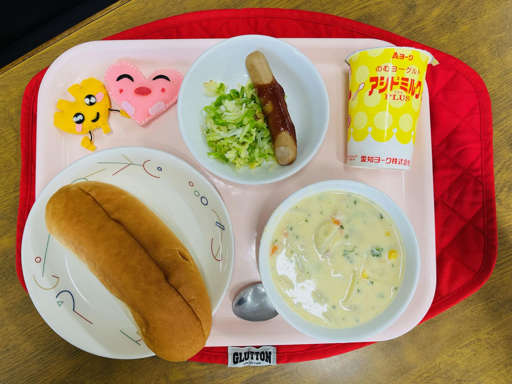 ピンクのトレーに給食が右上から反時計回りで発酵乳、ウインナーとキャベツソテー、ハートの形をした黄色とピンク色の布製マスコット、コッペパン、コーンシチューと並べられている写真