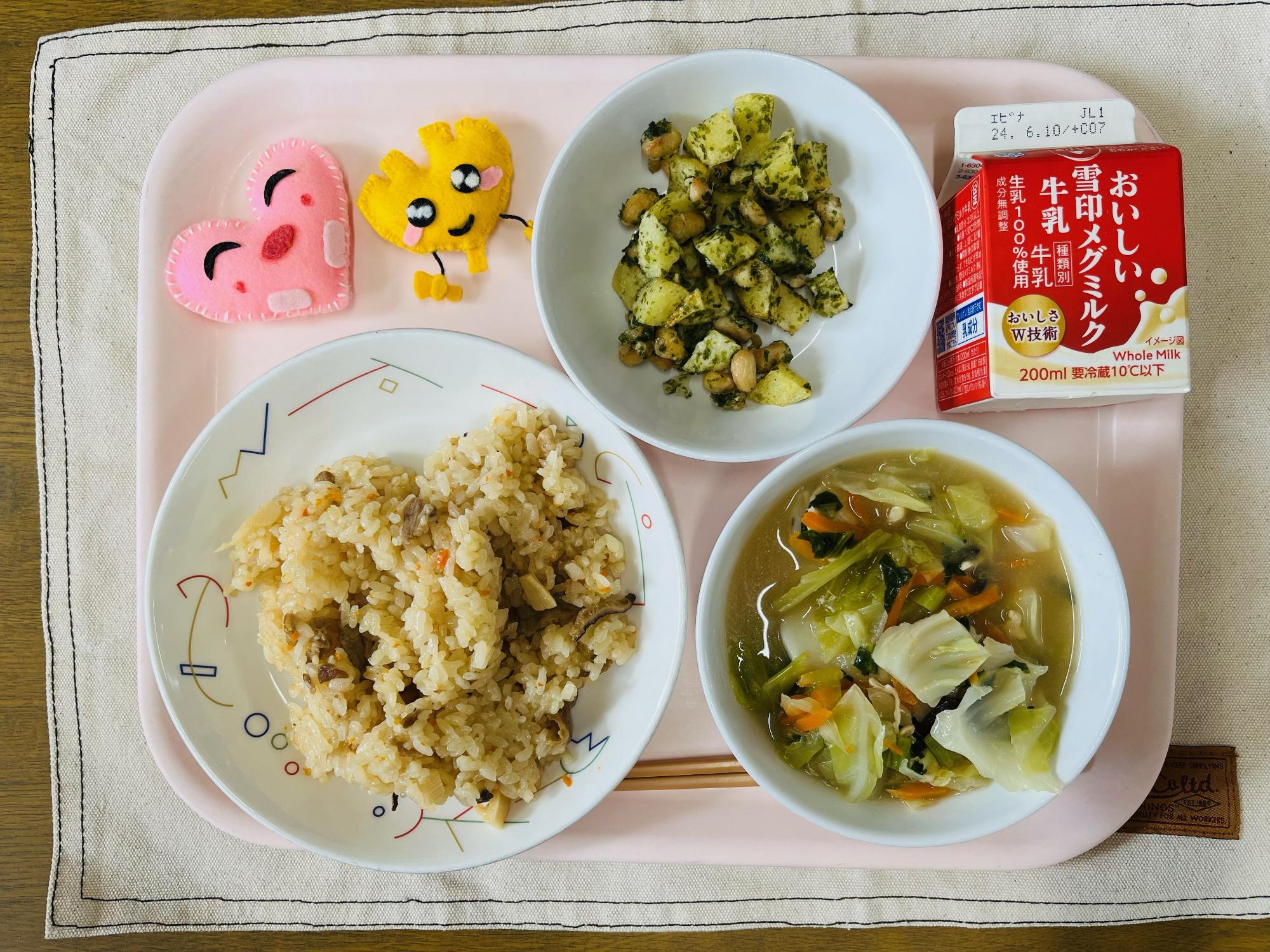 ピンクのトレーに給食が右上から反時計回りで牛乳、青のりポテトビーンズ、ハートの形をした黄色とピンク色の布製マスコット、中華風炊き込みご飯、トックスープと並べられている写真