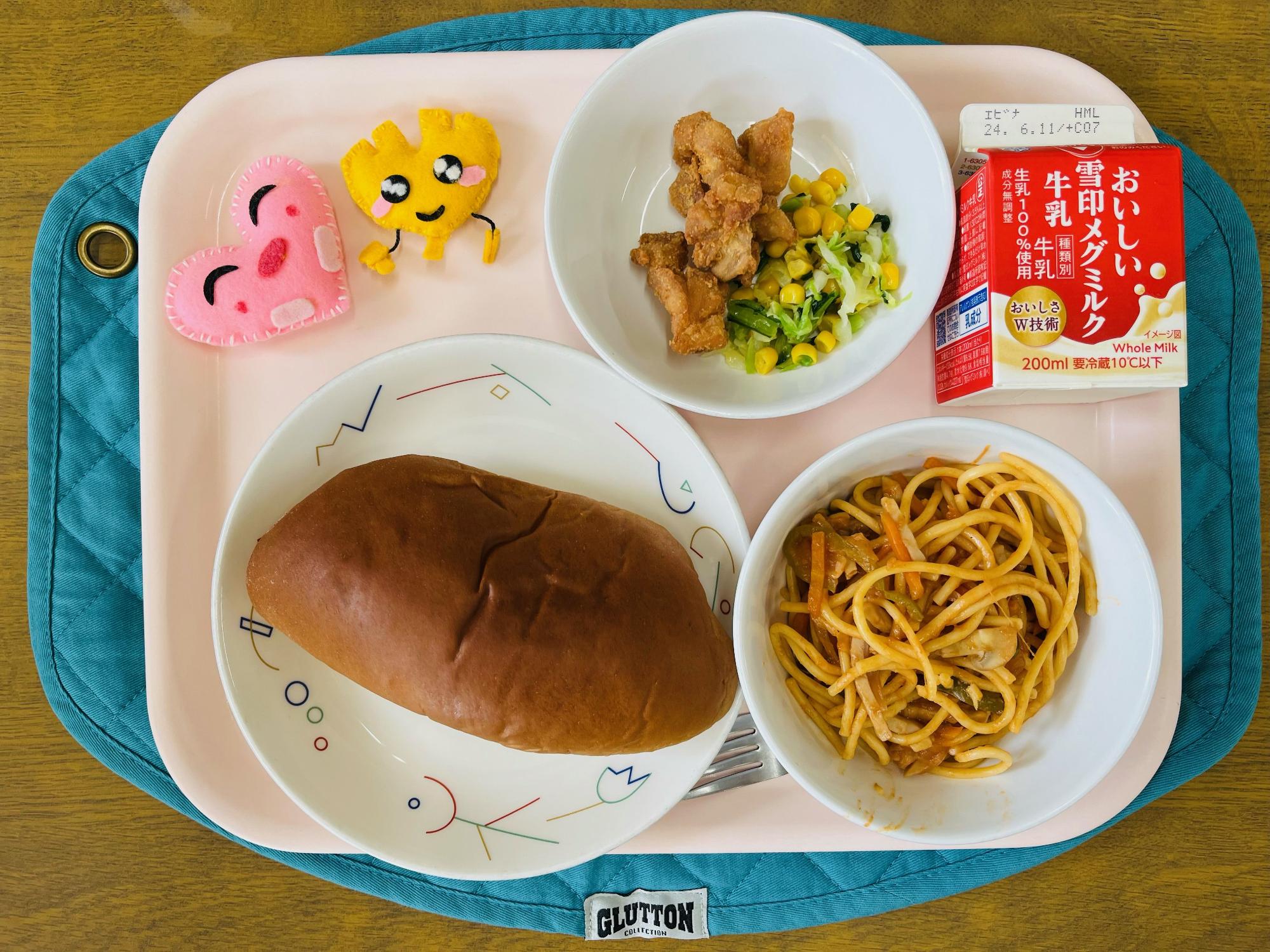ピンクのトレーに給食が右上から反時計回りで牛乳、鶏肉のからあげとミックスソテー、ハートの形をした黄色とピンク色の布製マスコット、くろパン、スパゲッティナポリタンと並べられている写真