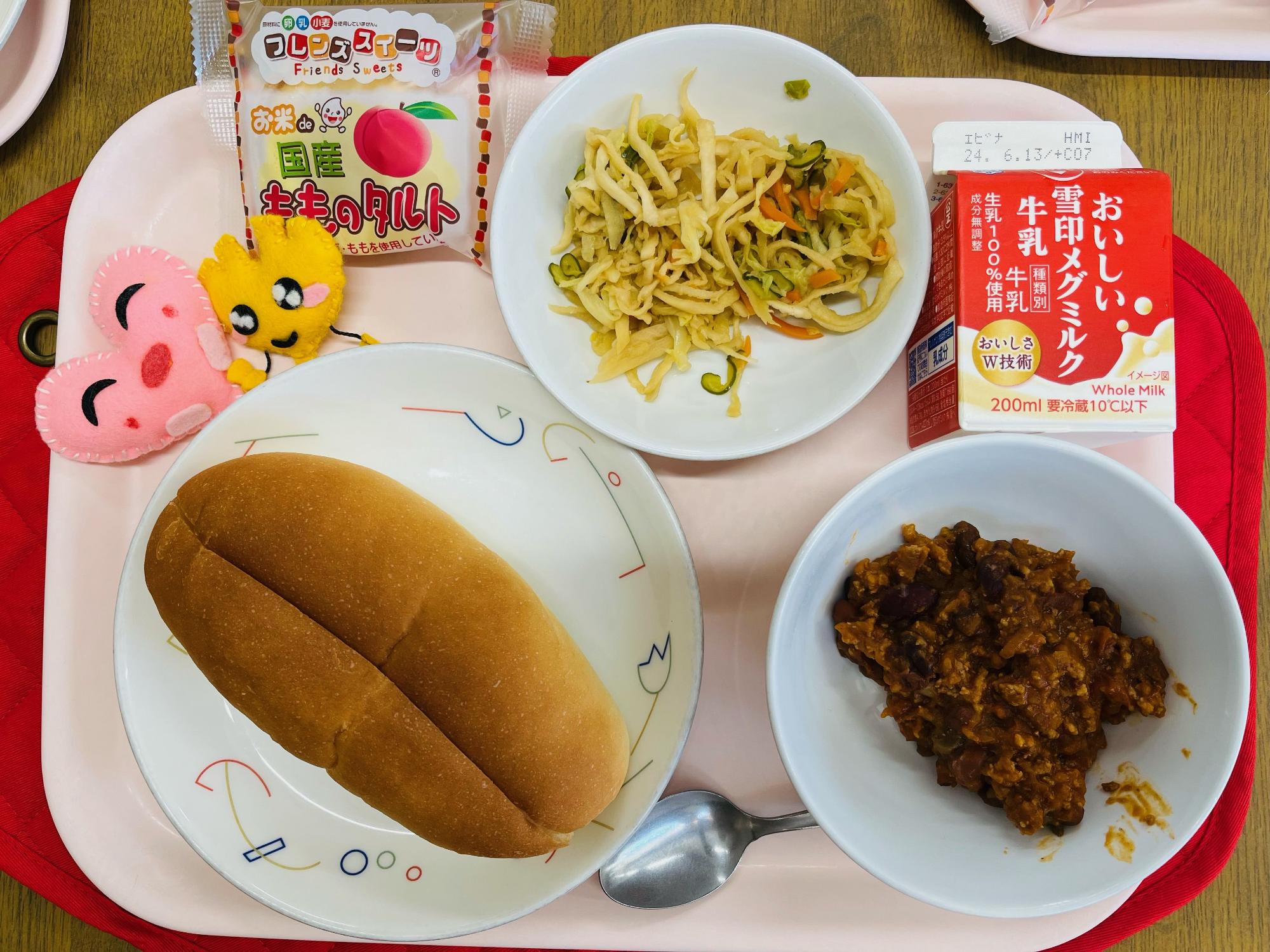 ピンクのトレーに給食が右上から反時計回りで牛乳、ハリハリサラダ、ももタルト、ハートの形をした黄色とピンク色の布製マスコット、ソフトフランスパン、チリコンカンと並べられている写真