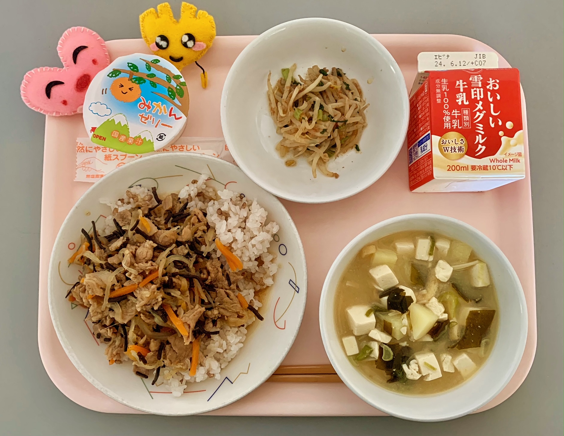 ピンクのトレーに給食が右上から反時計回りで牛乳、もやしのおかかあえ、みかんゼリー、ハートの形をした黄色とピンク色の布製マスコット、豚丼、みそしると並べられている写真