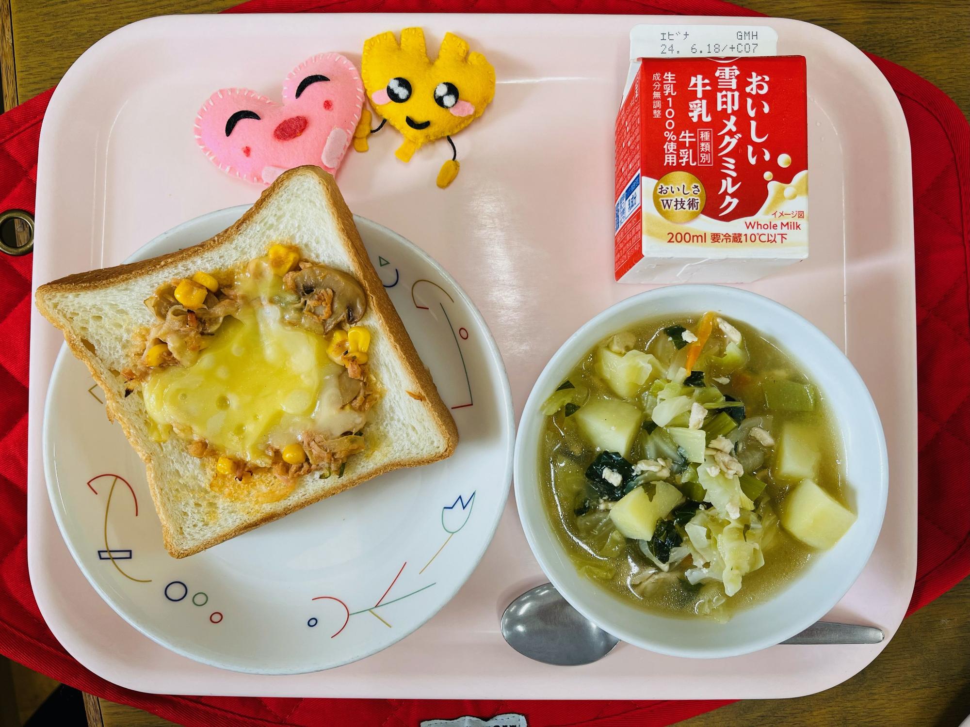 ピンクのトレーに給食が右上から反時計回りで牛乳、ハートの形をした黄色とピンク色の布製マスコット、ツナピザトースト、野菜のスープ煮と並べられている写真