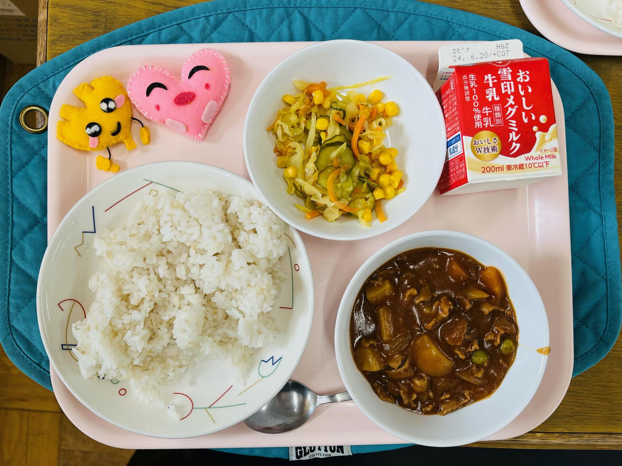 ピンクのトレーに給食が右上から反時計回りで牛乳、コーンサラダ、ハートの形をした黄色とピンク色の布製マスコット、むぎいりごはん、ハヤシライスと並べられている写真