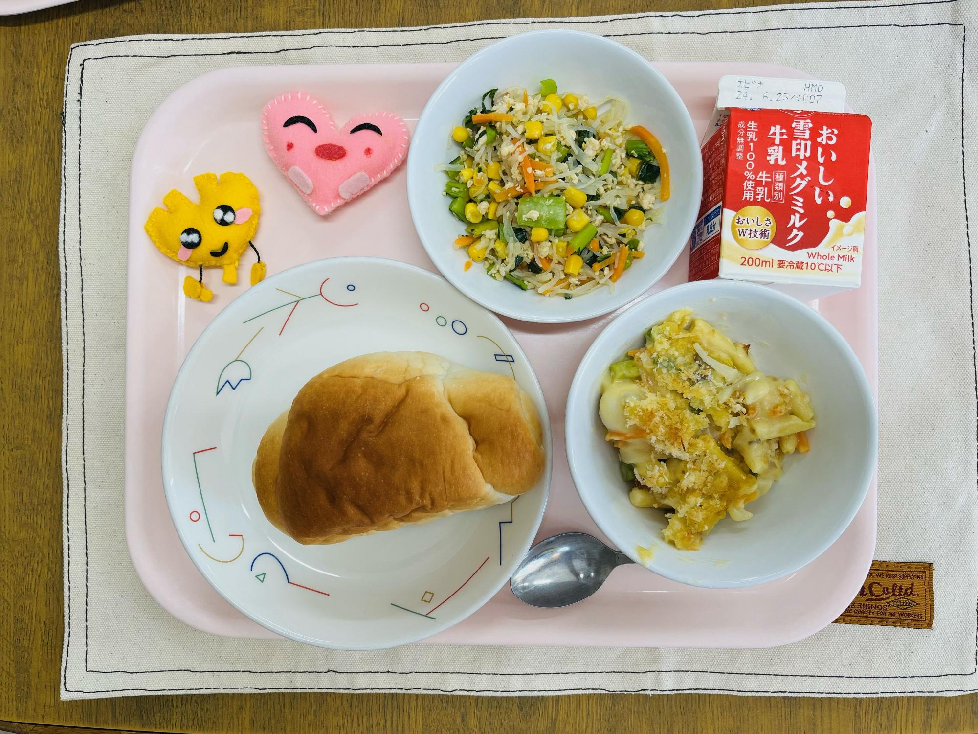 ピンクのトレーに給食が右上から反時計回りで牛乳、しらたきソテー、ハートの形をした黄色とピンク色の布製マスコット、こめこロールパン、アスパラガズとうずらたまごの豆乳グラタンと並べられている写真