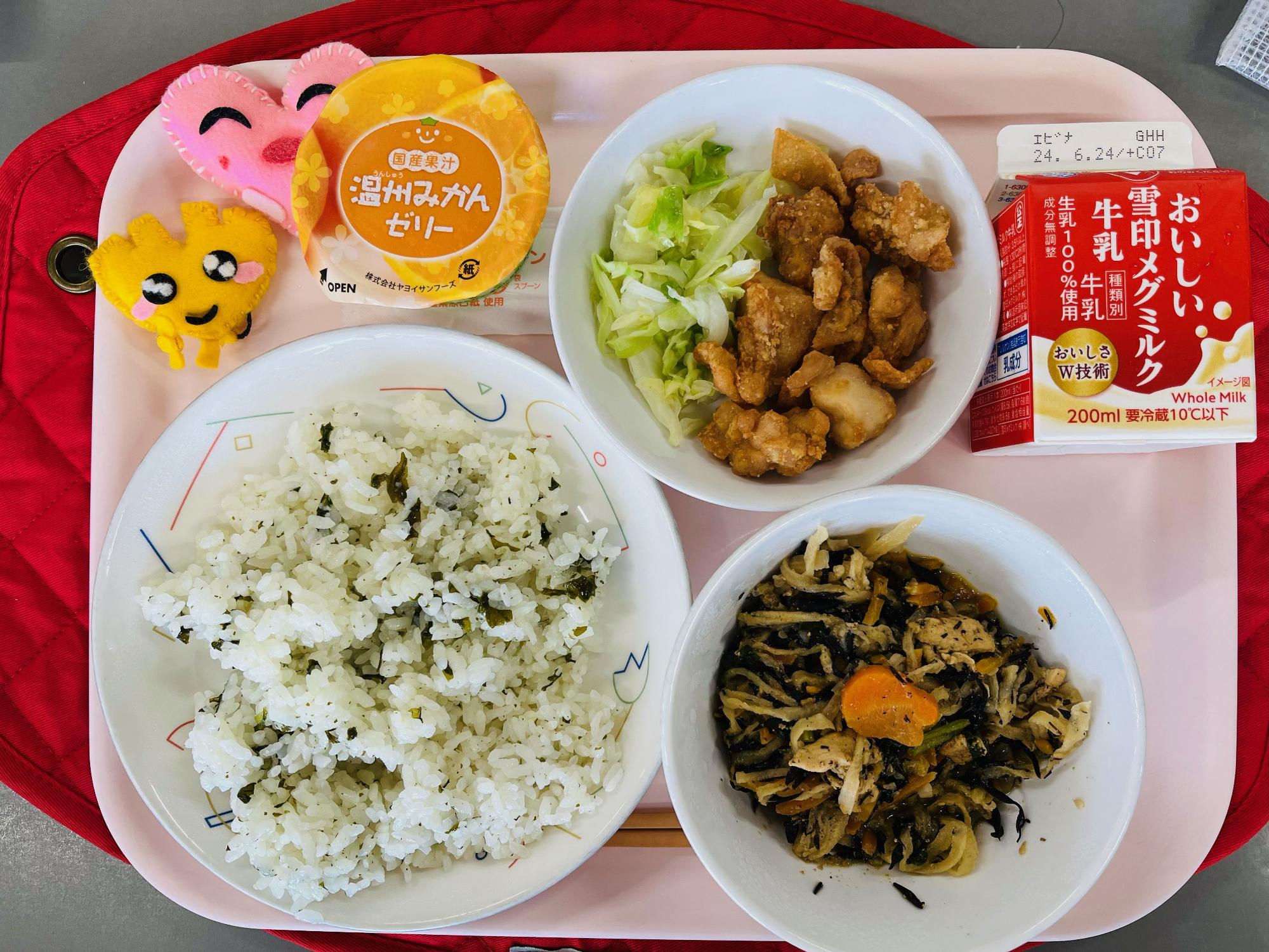 ピンクのトレーに給食が右上から反時計回りで牛乳、鶏肉のからあげとキャベツソテー、みかんゼリー、ハートの形をした黄色とピンク色の布製マスコット、わかめごはん、切り干し大根のミルク煮と並べられている写真