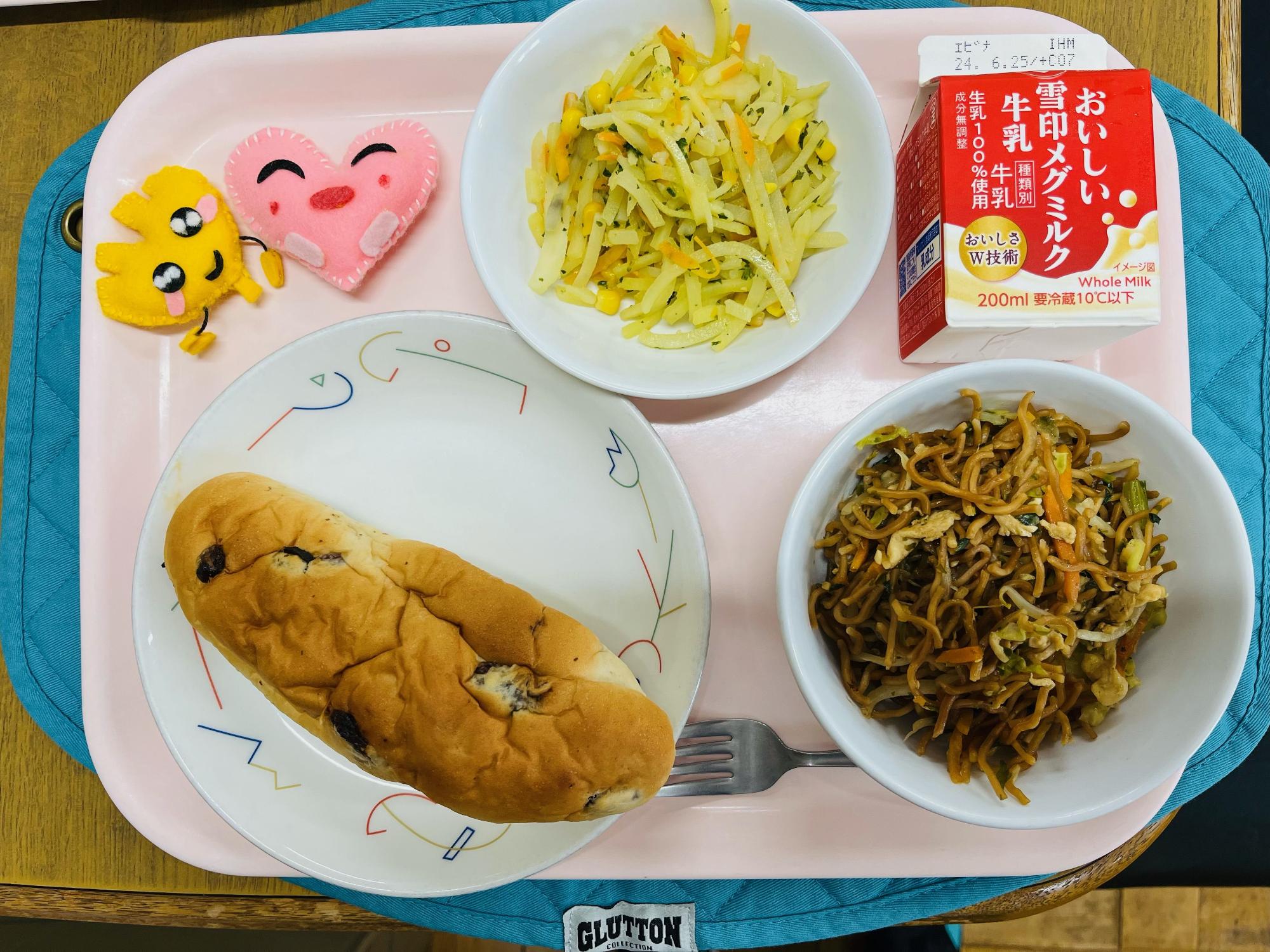 ピンクのトレーに給食が右上から反時計回りで牛乳、じゃがいものソテー、ハートの形をした黄色とピンク色の布製マスコット、ぶどうパン、ソース焼きそばと並べられている写真