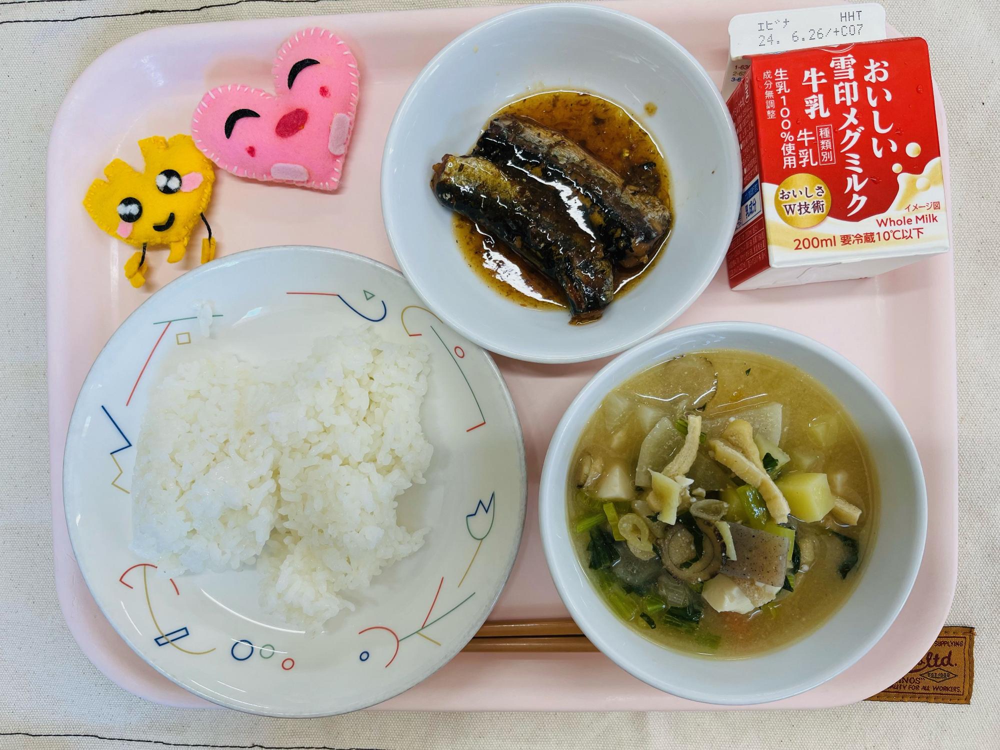 ピンクのトレーに給食が右上から反時計回りで牛乳、いわしの梅煮、ハートの形をした黄色とピンク色の布製マスコット、ごはん、みそけんちん汁と並べられている写真