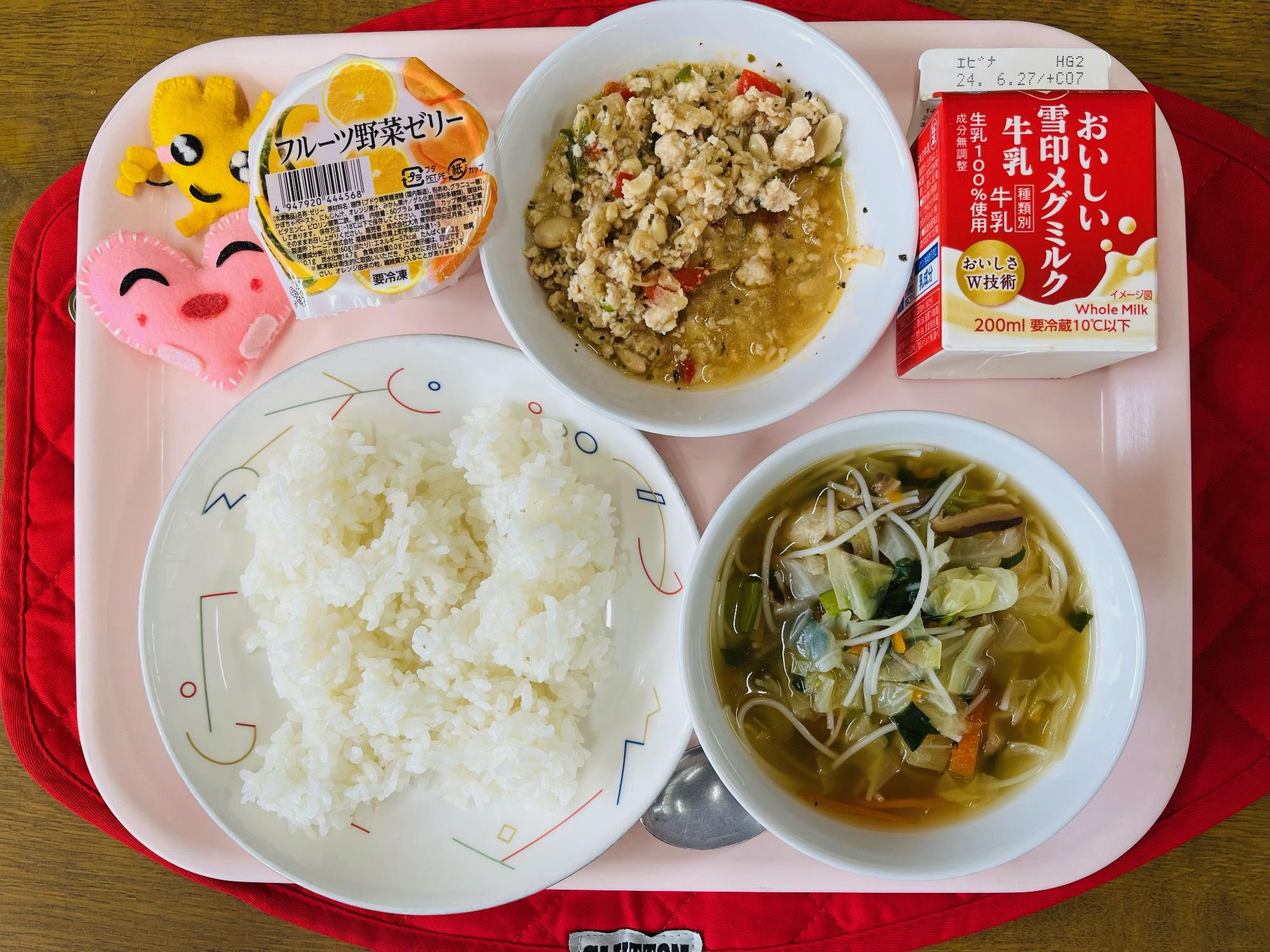 ピンクのトレーに給食が右上から反時計回りで牛乳、ガパオライスの具、フルーツ野菜ゼリー、ハートの形をした黄色とピンク色の布製マスコット、ごはん、鶏団子スープと並べられている写真