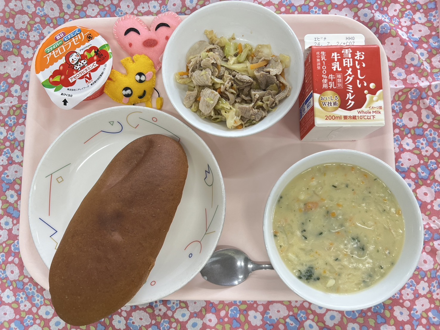 ピンクのトレーに給食が右上から反時計回りで牛乳、ゆでぶたサラダ、ハートの形をした黄色とピンク色の布製マスコット、アセロラゼリー、くろパン、ポテトスープと並べられている写真