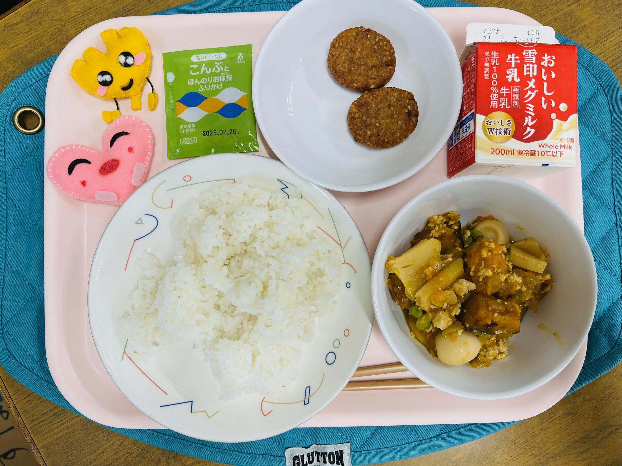 ピンクのトレーに給食が右上から反時計回りで牛乳、しろごまつくね、抹茶こんぶふりかけ、ハートの形をした黄色とピンク色の布製マスコット、ごはん、かぼちゃのそぼろ煮と並べられている写真