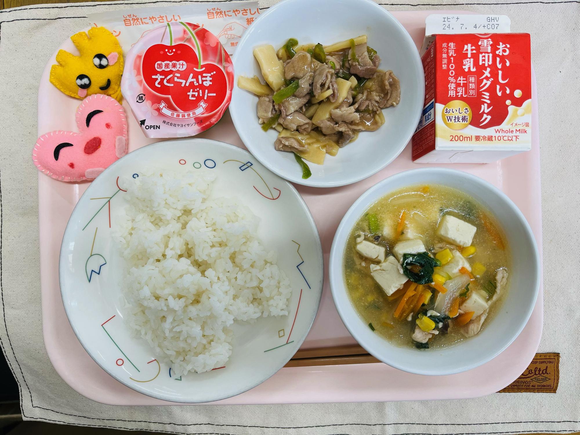 ピンクのトレーに給食が右上から反時計回りで牛乳、豚肉とピーマンの炒め物、さくらんぼゼリー、ハートの形をした黄色とピンク色の布製マスコット、ごはん、コーン中華スープと並べられている写真