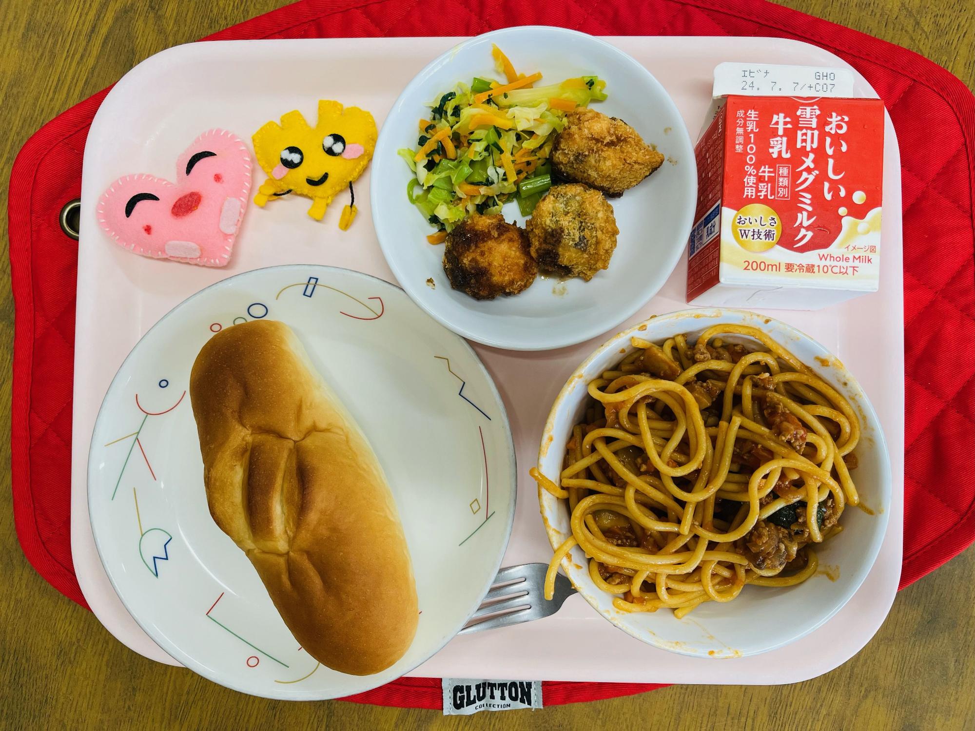 ピンクのトレーに給食が右上から反時計回りで牛乳、ミックスソテーとさけフライ、ハートの形をした黄色とピンク色の布製マスコット、ミルクパン、スッキーニのミートスパゲティと並べられている写真