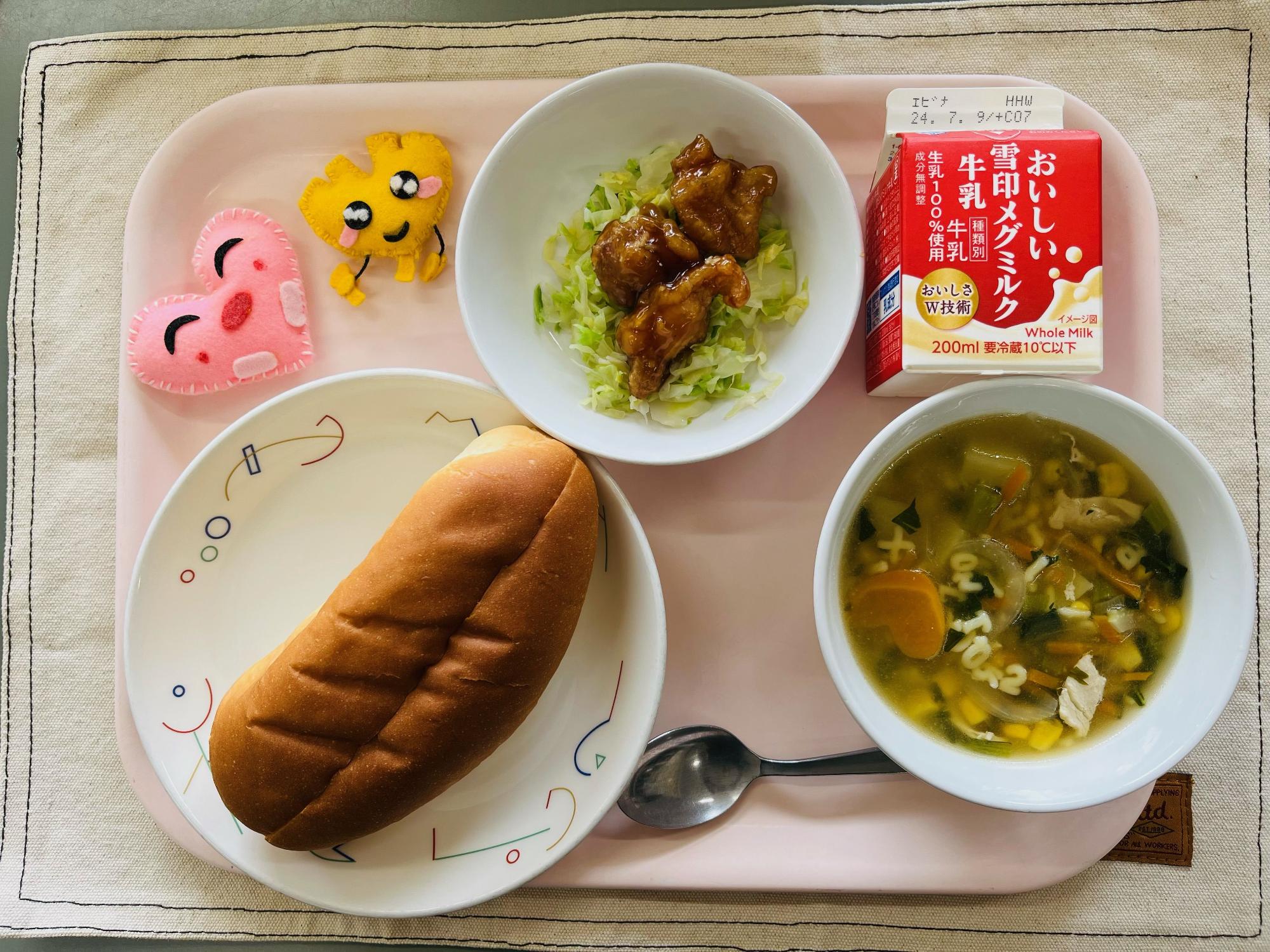 ピンクのトレーに給食が右上から反時計回りで牛乳、照り焼きチキンと野菜、ハートの形をした黄色とピンク色の布製マスコット、サンドパン、ABCスープと並べられている写真