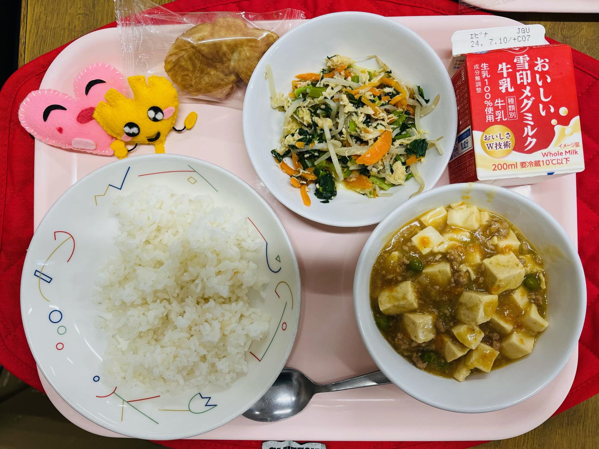 ピンクのトレーに給食が右上から反時計回りで牛乳、鶏肉と野菜のナムル、プチたいやき、ハートの形をした黄色とピンク色の布製マスコット、ごはん、マーボー豆腐と並べられている写真