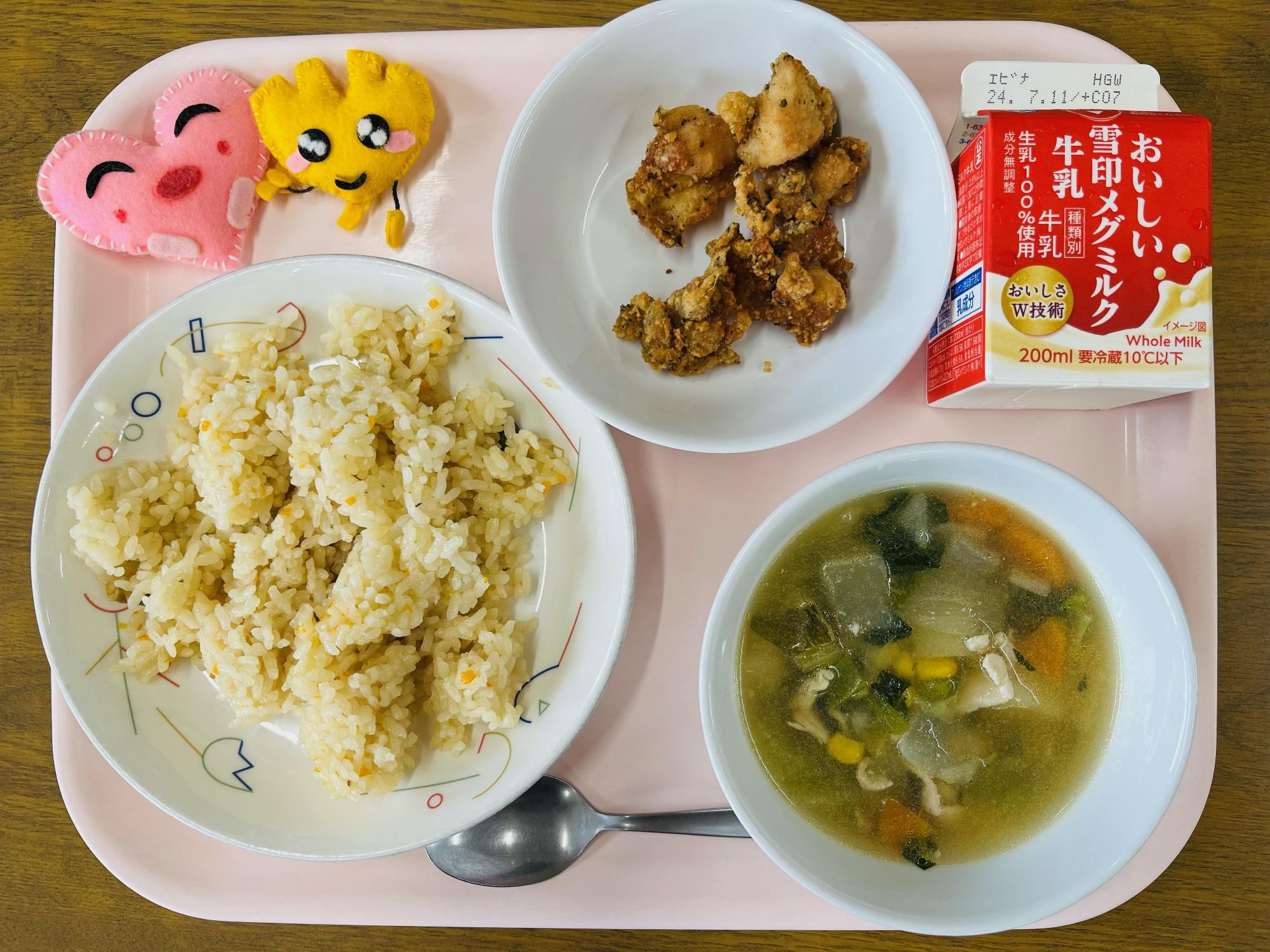 ピンクのトレーに給食が右上から反時計回りで牛乳、鶏肉のハーブ揚げ、ハートの形をした黄色とピンク色の布製マスコット、ガーリックライス、野菜スープと並べられている写真