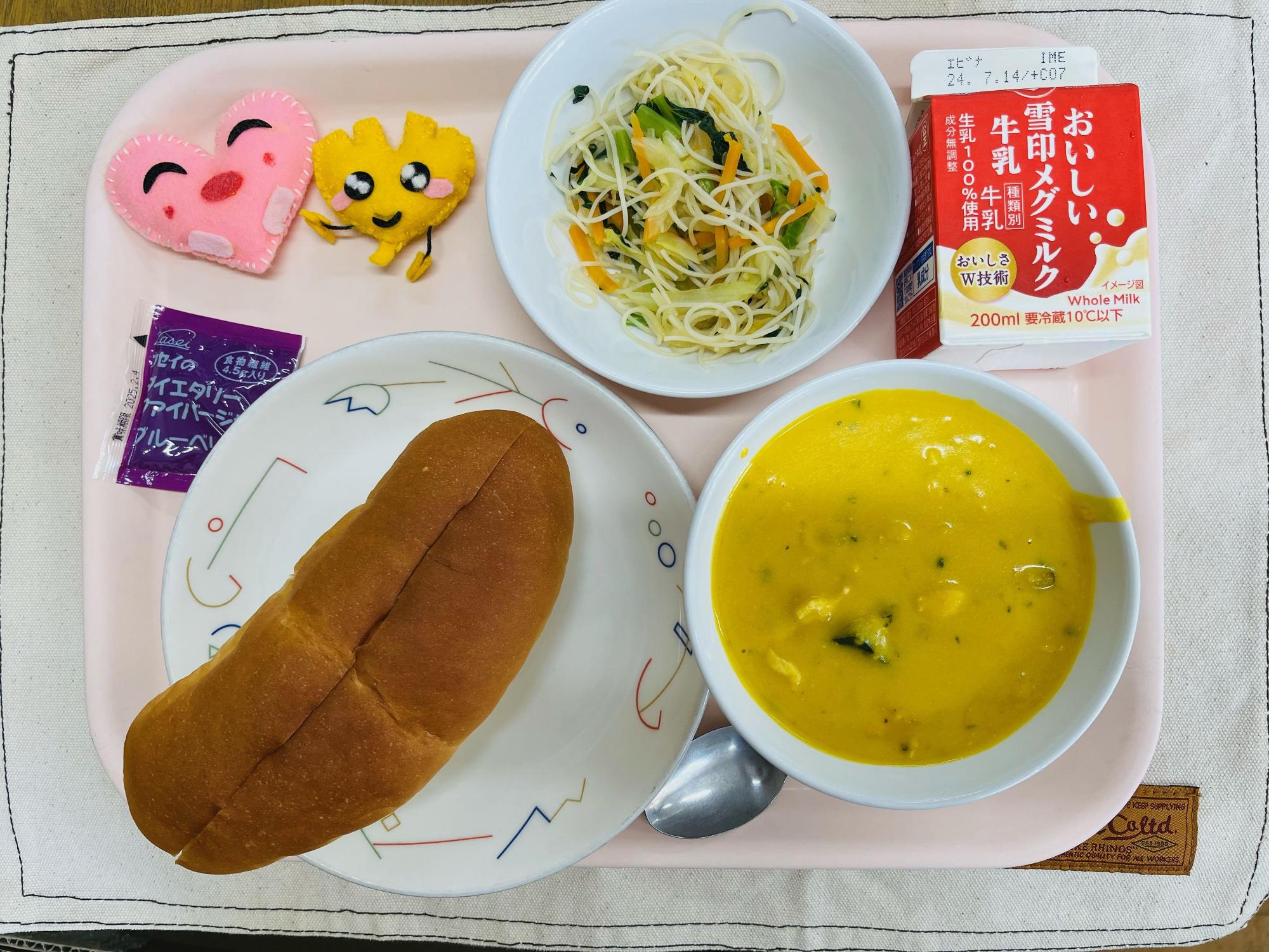ピンクのトレーに給食が右上から反時計回りで牛乳、ビーフンソテー、ハートの形をした黄色とピンク色の布製マスコット、ブルーベリージャム、ソフトフランスパン、かぼちゃのクリームスープと並べられている写真