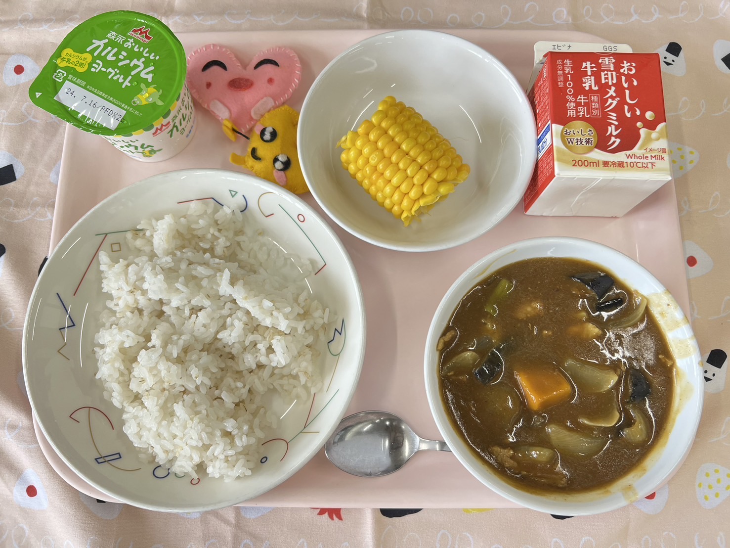 ピンクのトレーに給食が右上から反時計回りで牛乳、とうもろこし、ハートの形をした黄色とピンク色の布製マスコット、ヨーグルト、ごはん、夏野菜のカレーと並べられている写真
