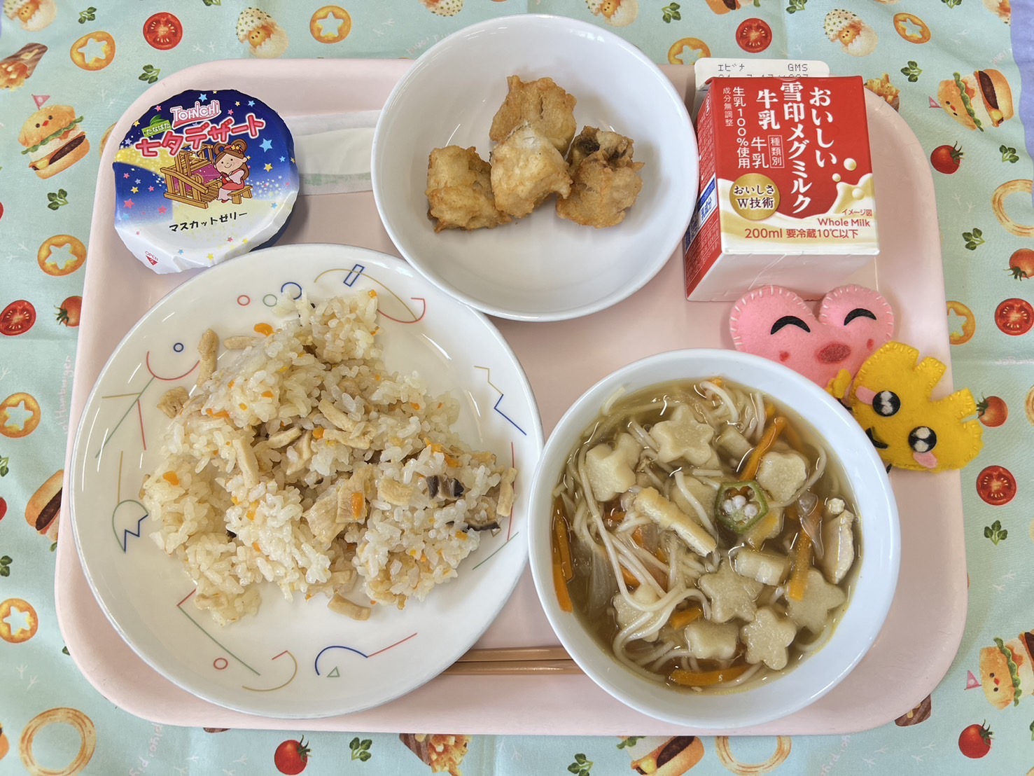 ピンクのトレーに給食が右上から反時計回りで牛乳、さかなの唐揚げ、七夕ゼリー、かしわずし、七夕汁、ハートの形をした黄色とピンク色の布製マスコットと並べられている写真