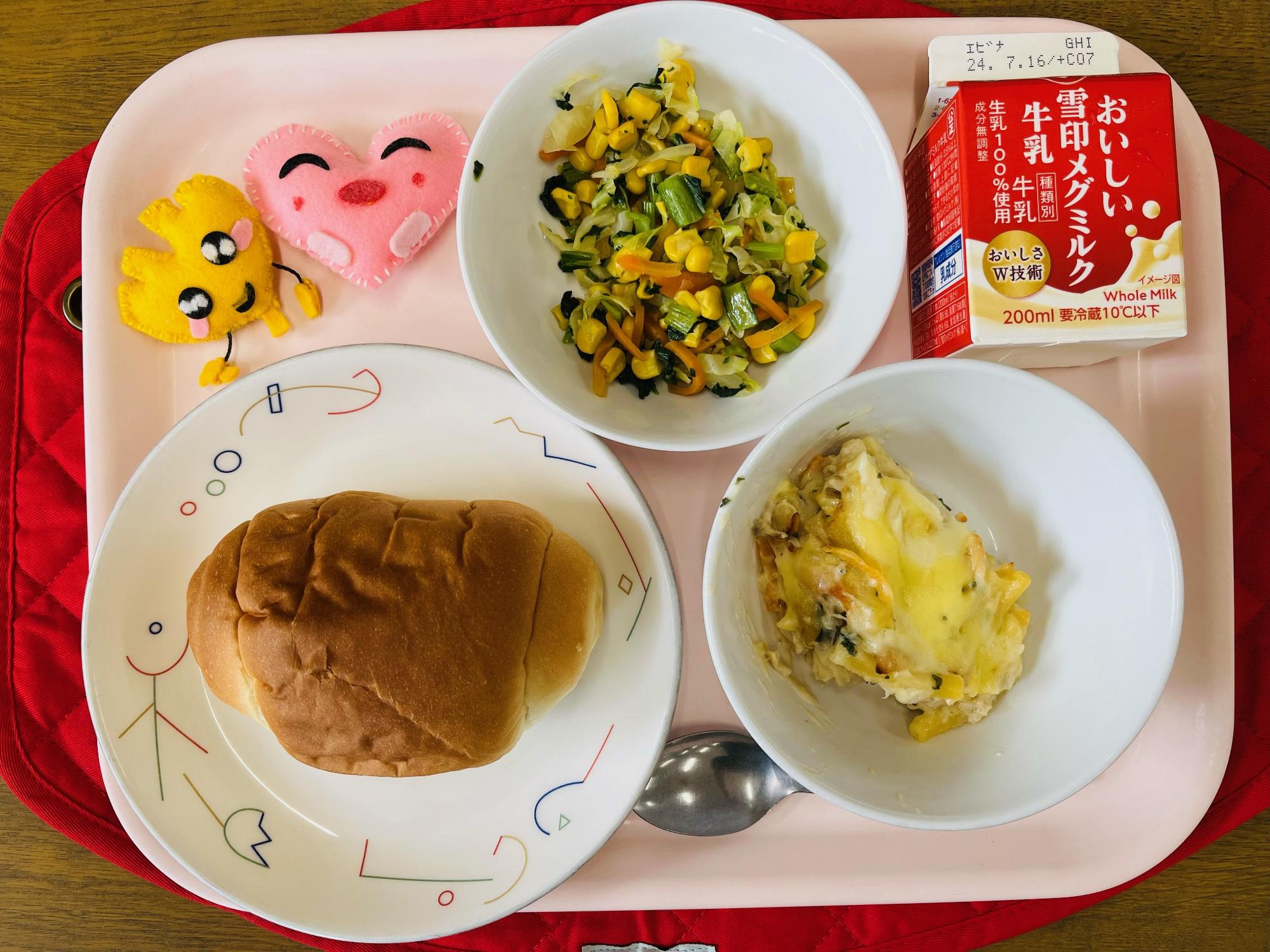 ピンクのトレーに給食が右上から反時計回りで牛乳、野菜ソテー、ハートの形をした黄色とピンク色の布製マスコット、ロールパン、マカロニグラタンと並べられている写真